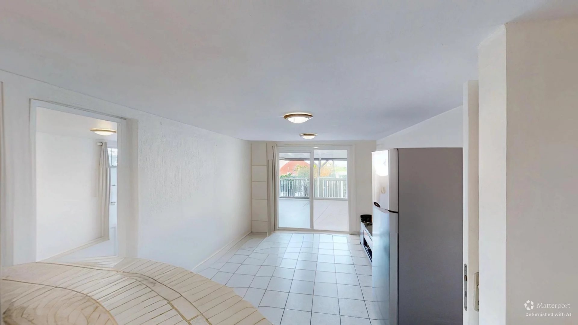 Bright white, open-plan living area with tiled floor, large sliding glass door to a balcony, and a refrigerator against the right wall.