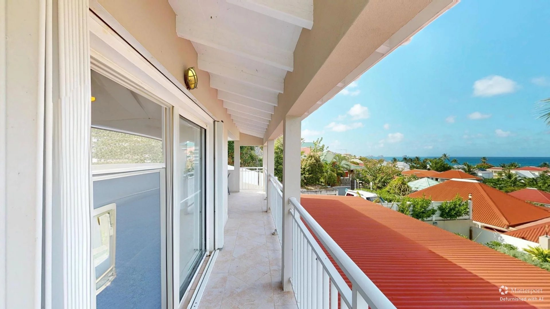 Beachside balcony with sliding glass doors, white railing, and a view of orange rooftops and the ocean.