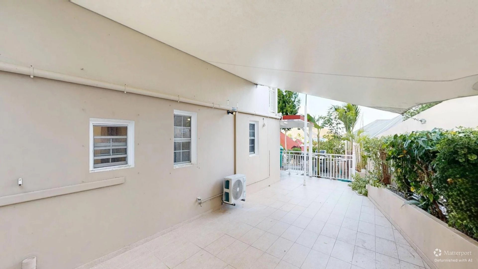 A spacious beige patio with tiled floor, two white-framed windows, an outdoor AC unit, and a covered shaded area leading to a railing and garden beyond.