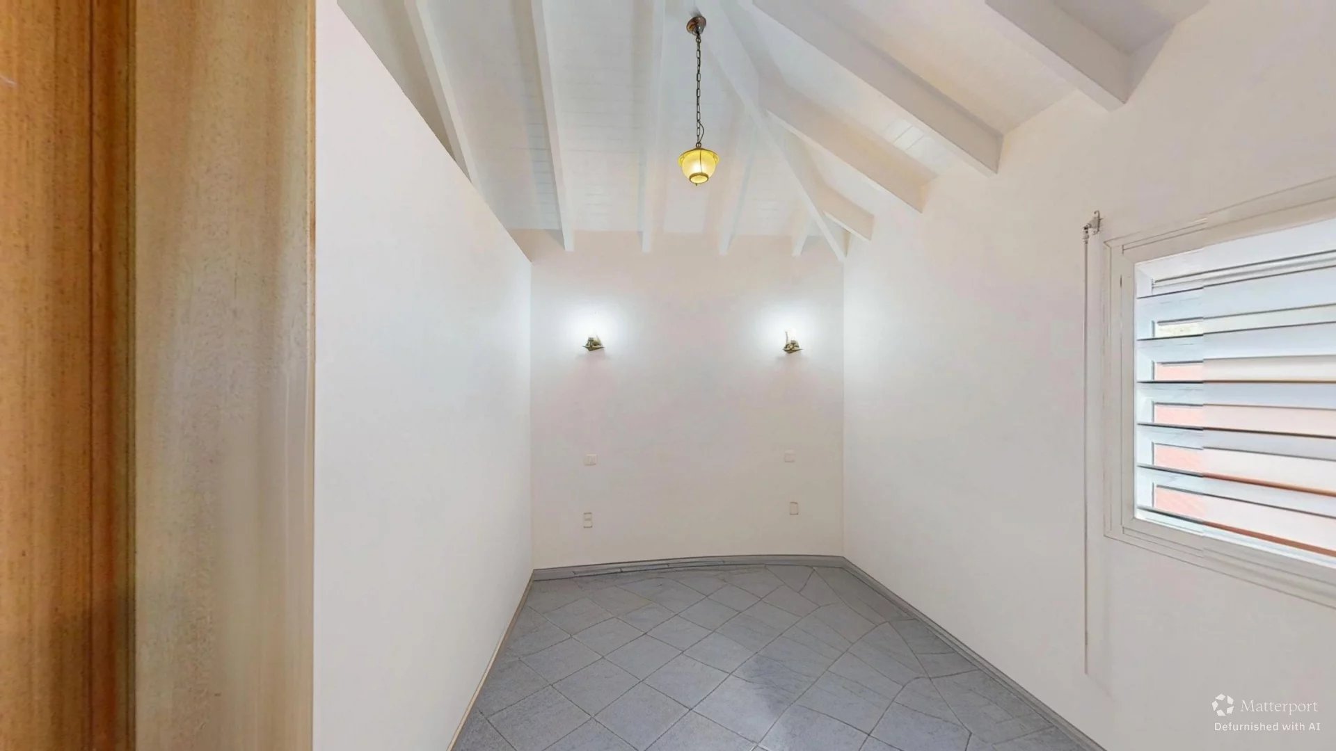 Empty white-walled room with exposed sloped ceiling beams, a hanging pendant light, and two wall sconces; window with blinds on the right.