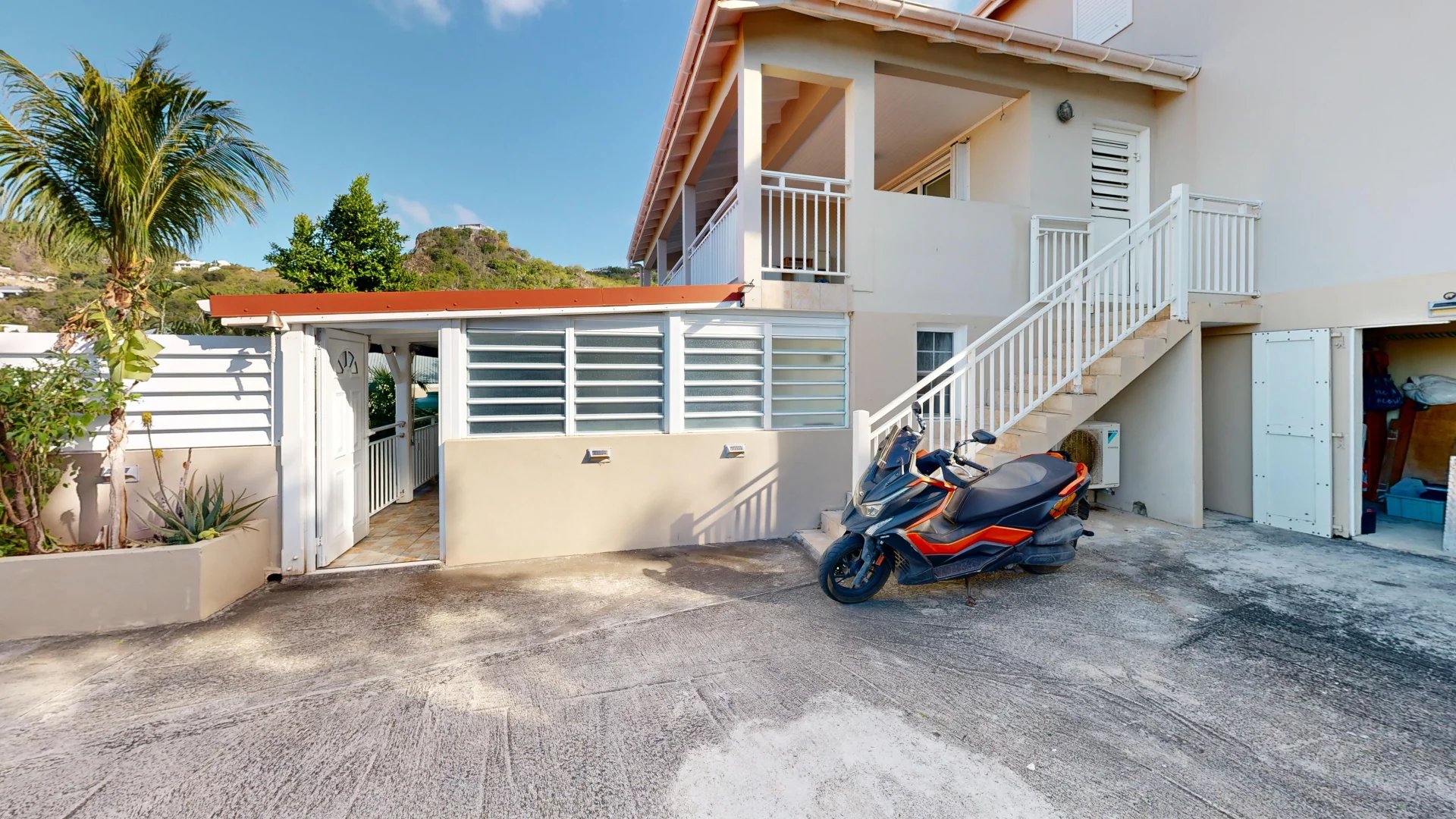 Beige two-story building with white railings and an external staircase; a black and orange scooter parked on the concrete driveway.