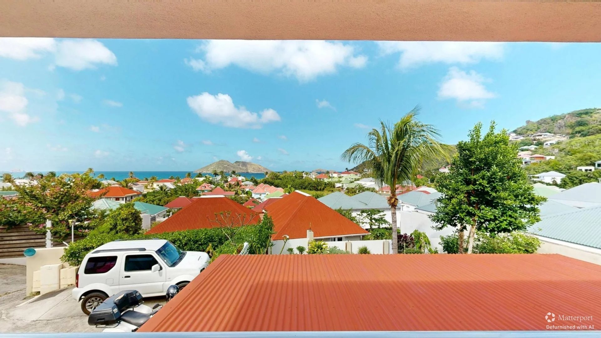 Coastal neighborhood with colorful red-tiled roofs, palm trees, and a bright blue sky; a white van and motorcycle are in the foreground near an orange roof.