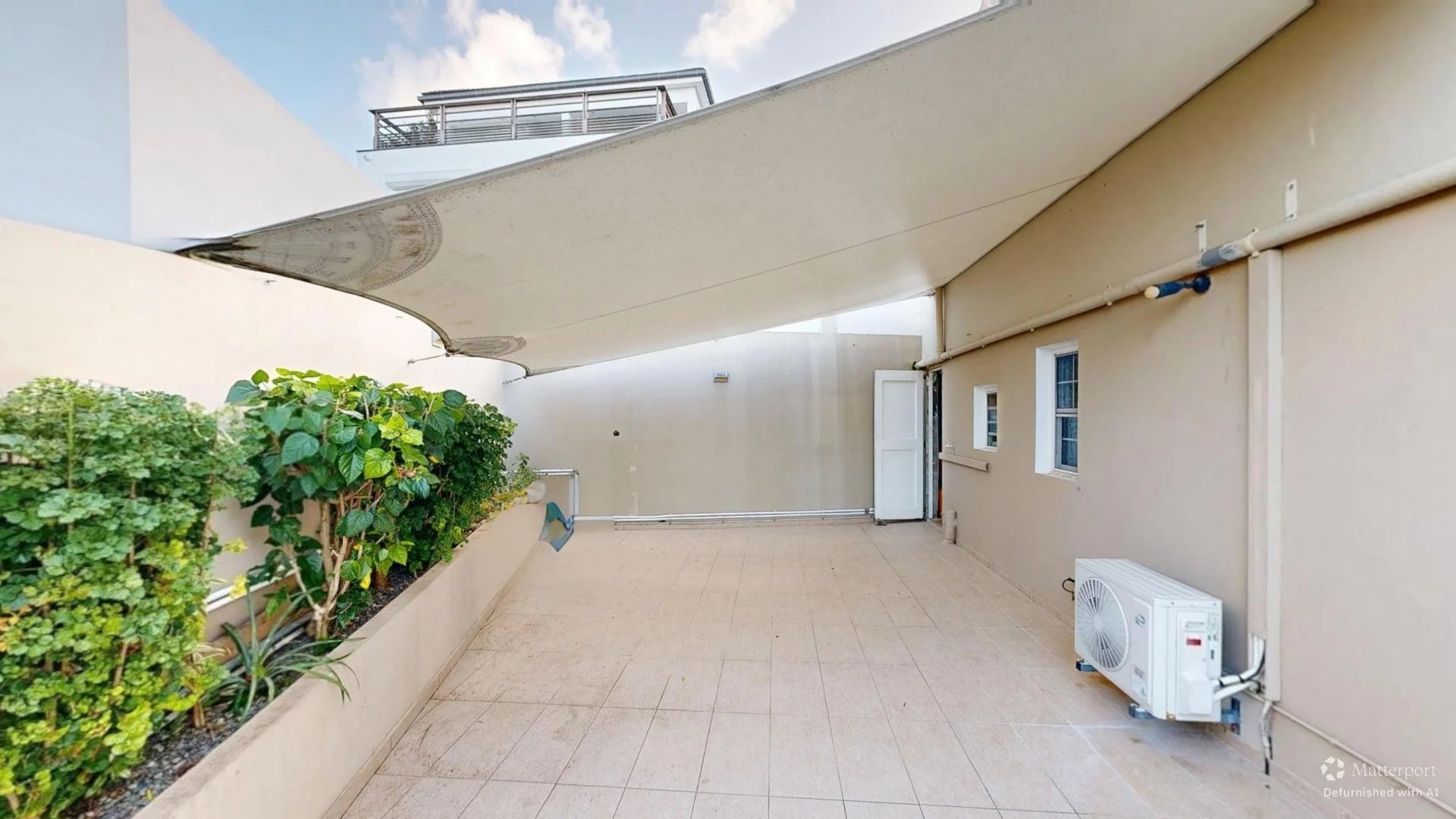 Rooftop terrace with a large diagonal shade sail, beige walls, tiled floor, and potted plants along a planter on the left.