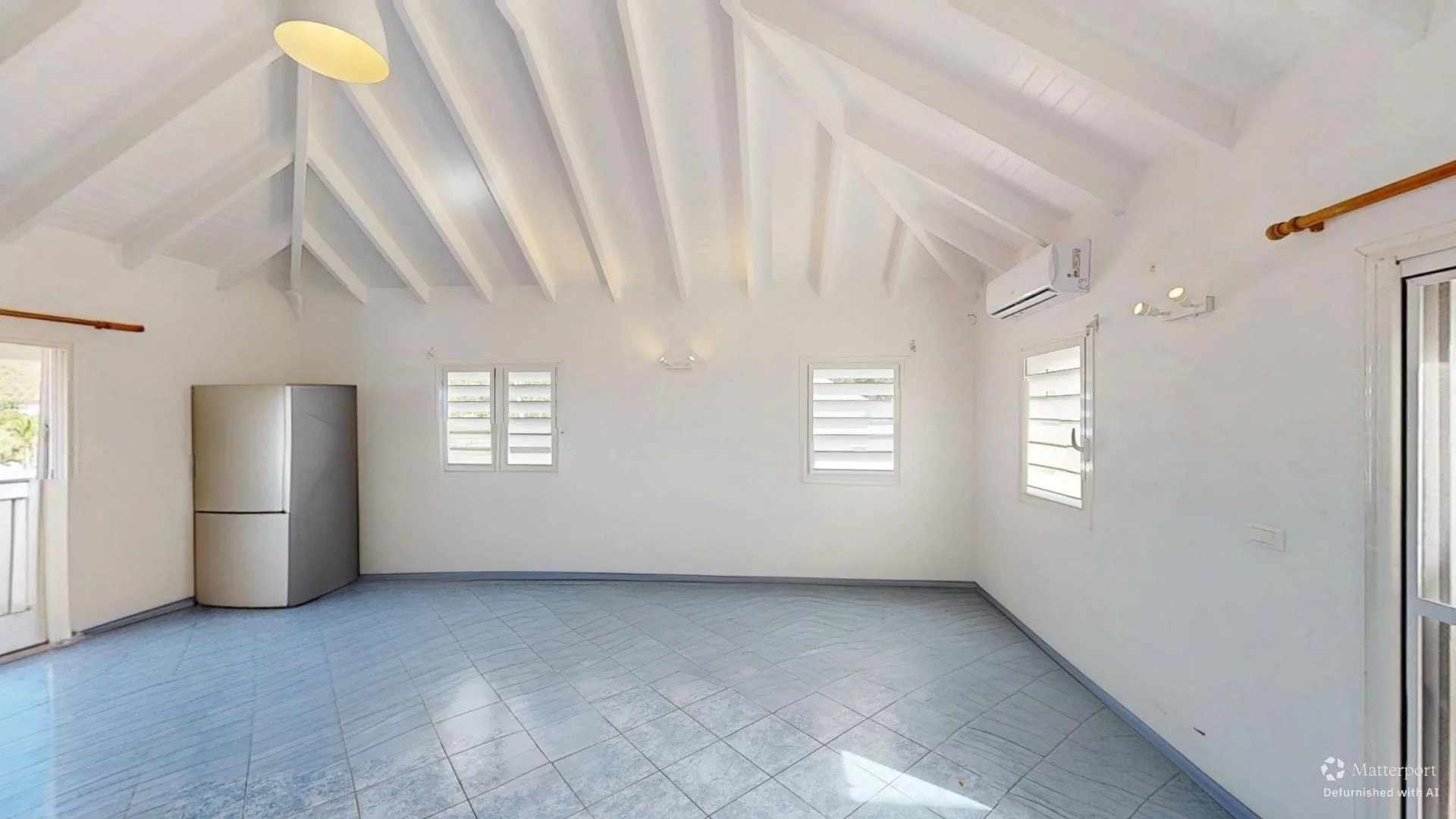 Bright white loft-style room with a vaulted ceiling, exposed white beams, and a blue tiled floor; a fridge sits in the corner.