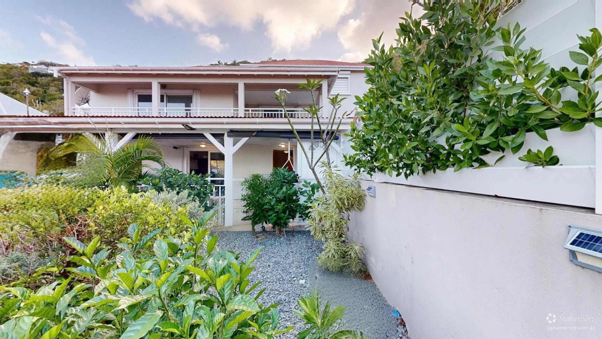 Front of a two-story house with a white railing, covered porch, and a gravel pathway flanked by lush tropical plants.