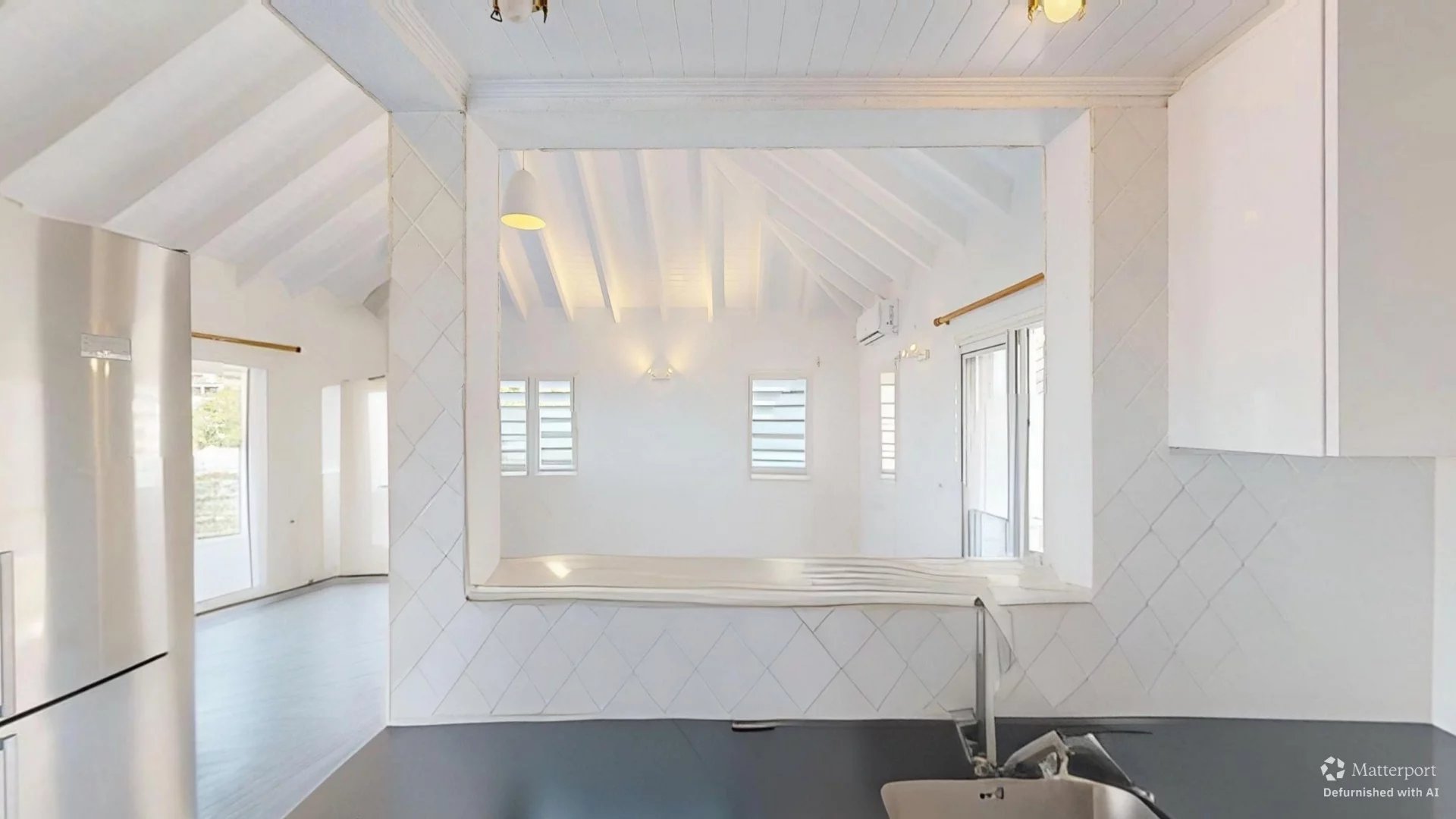 Bright white kitchen with a large passthrough window to a sunlit living area, stainless fridge, and dark countertop with a sink.