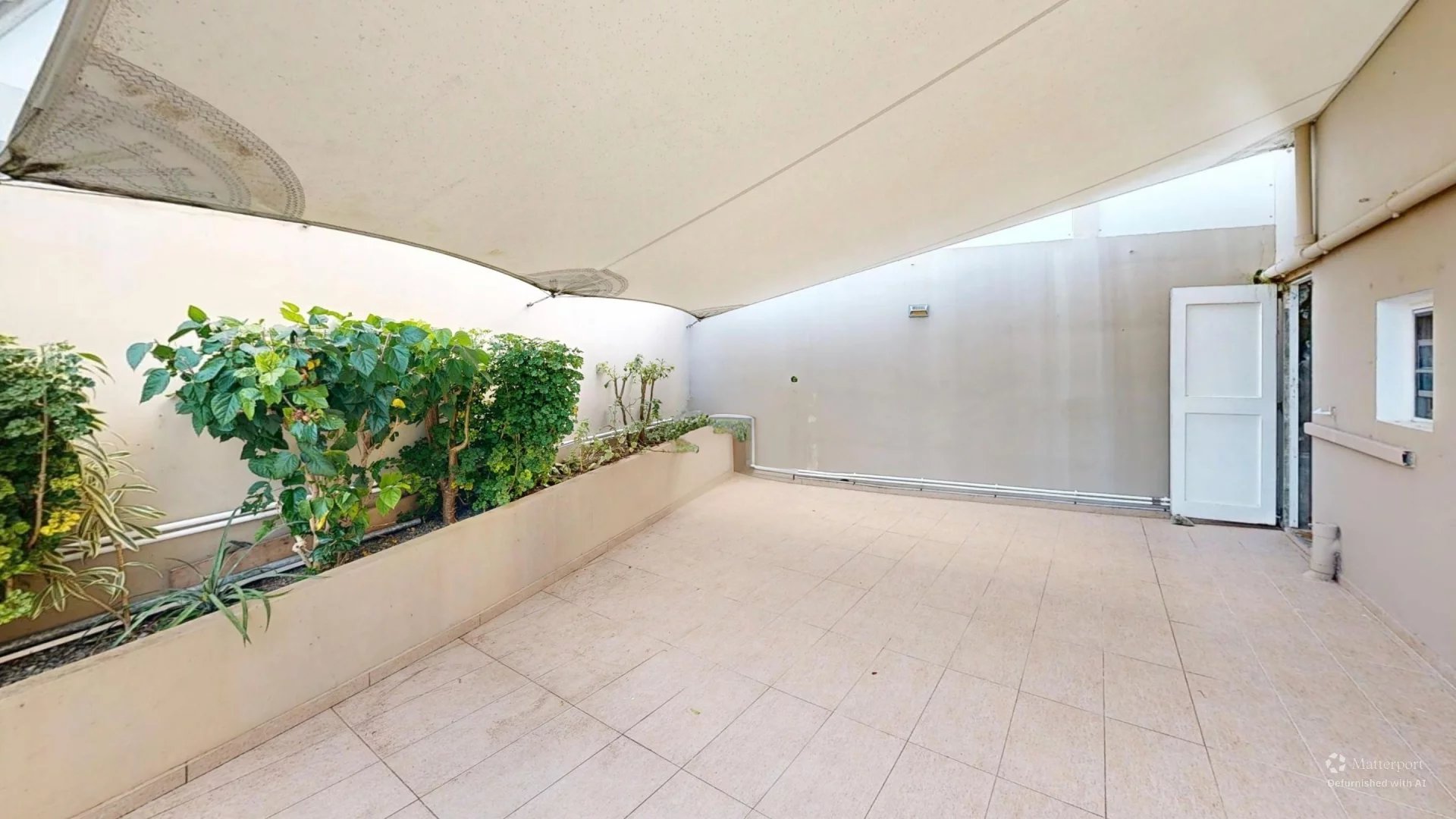 Open-air patio with beige tiled floor, a large white shade sail overhead, and a row of green plants in a raised planter along the left wall.