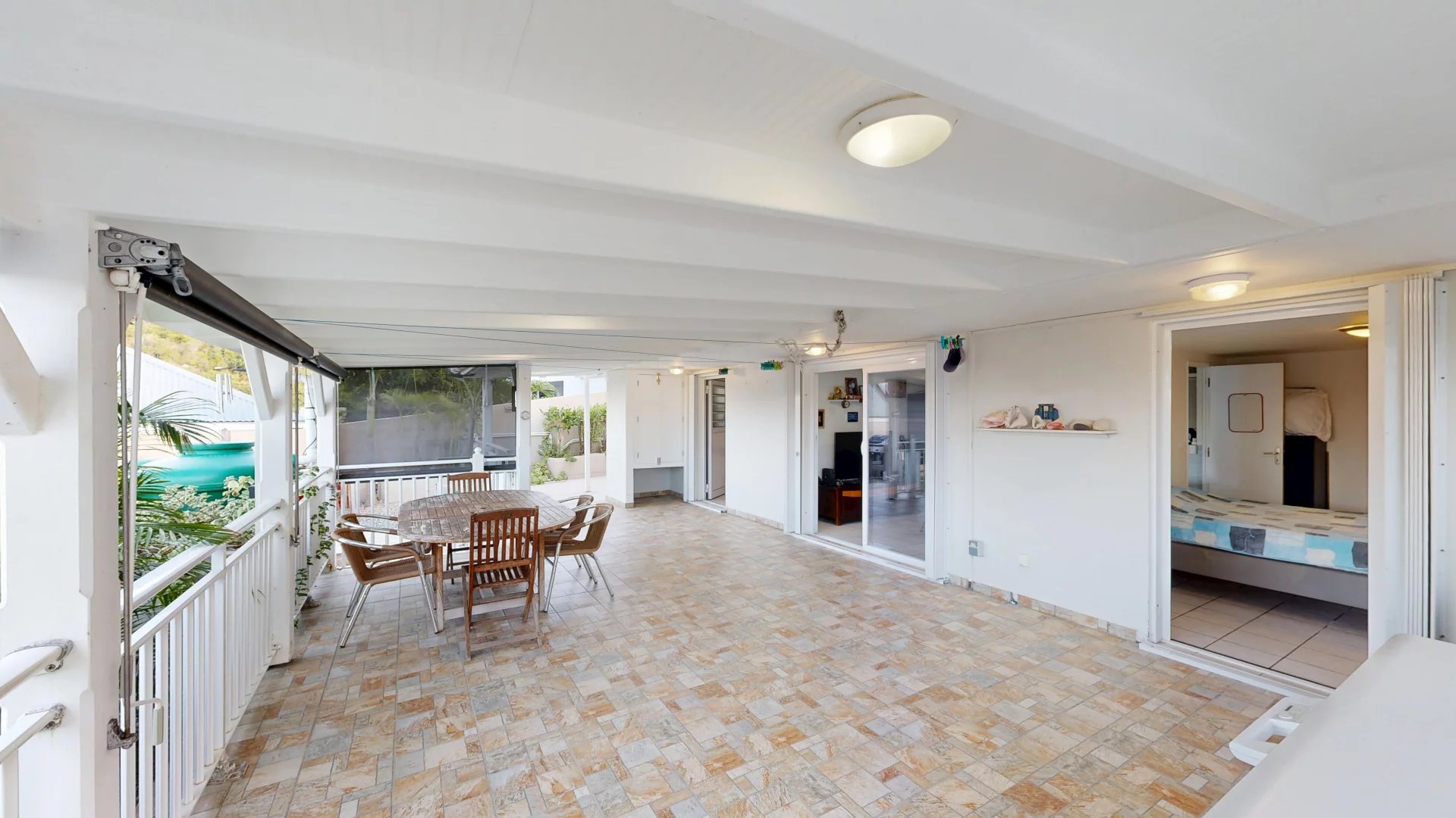 Covered patio with a round wooden table and six chairs, sliding doors to an interior bedroom, and white walls/orange-tiled floor.