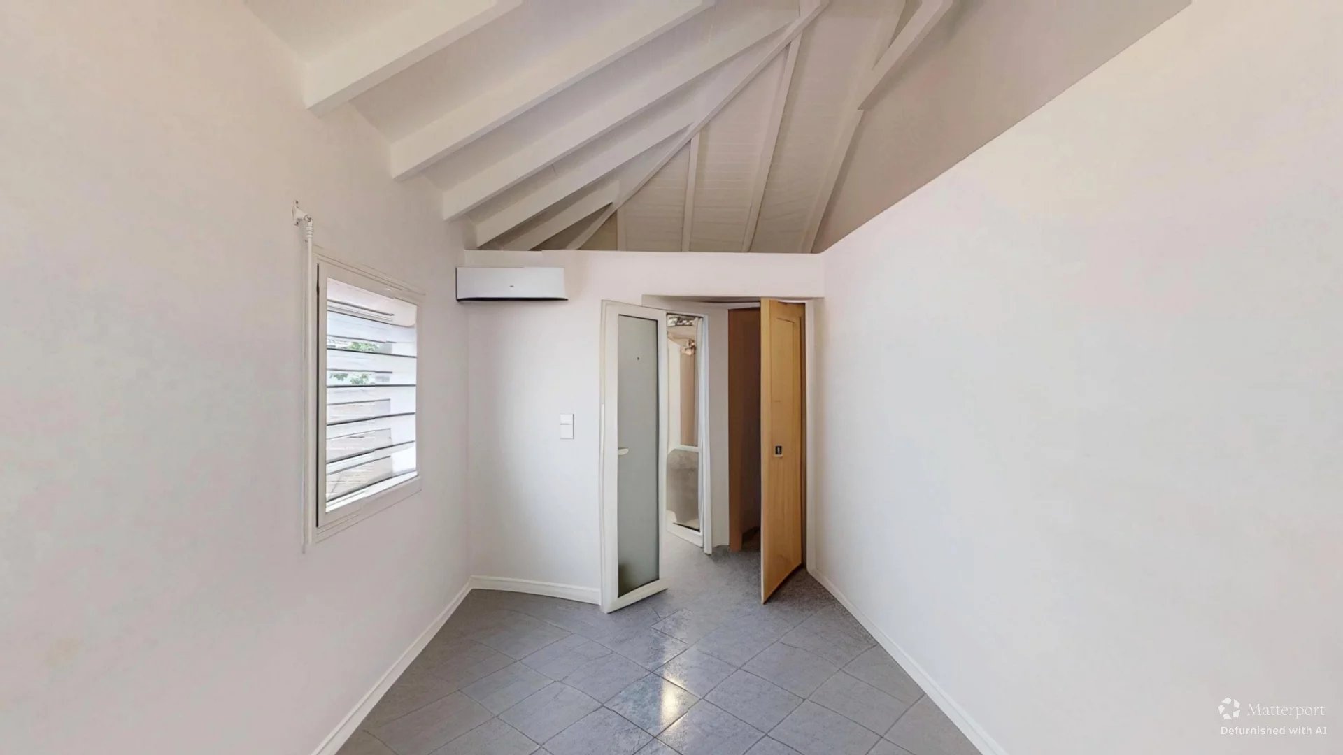 Small white room with a slanted ceiling, window with louvers, wall-mounted air conditioner, and a doorway leading to a bathroom/shower area.