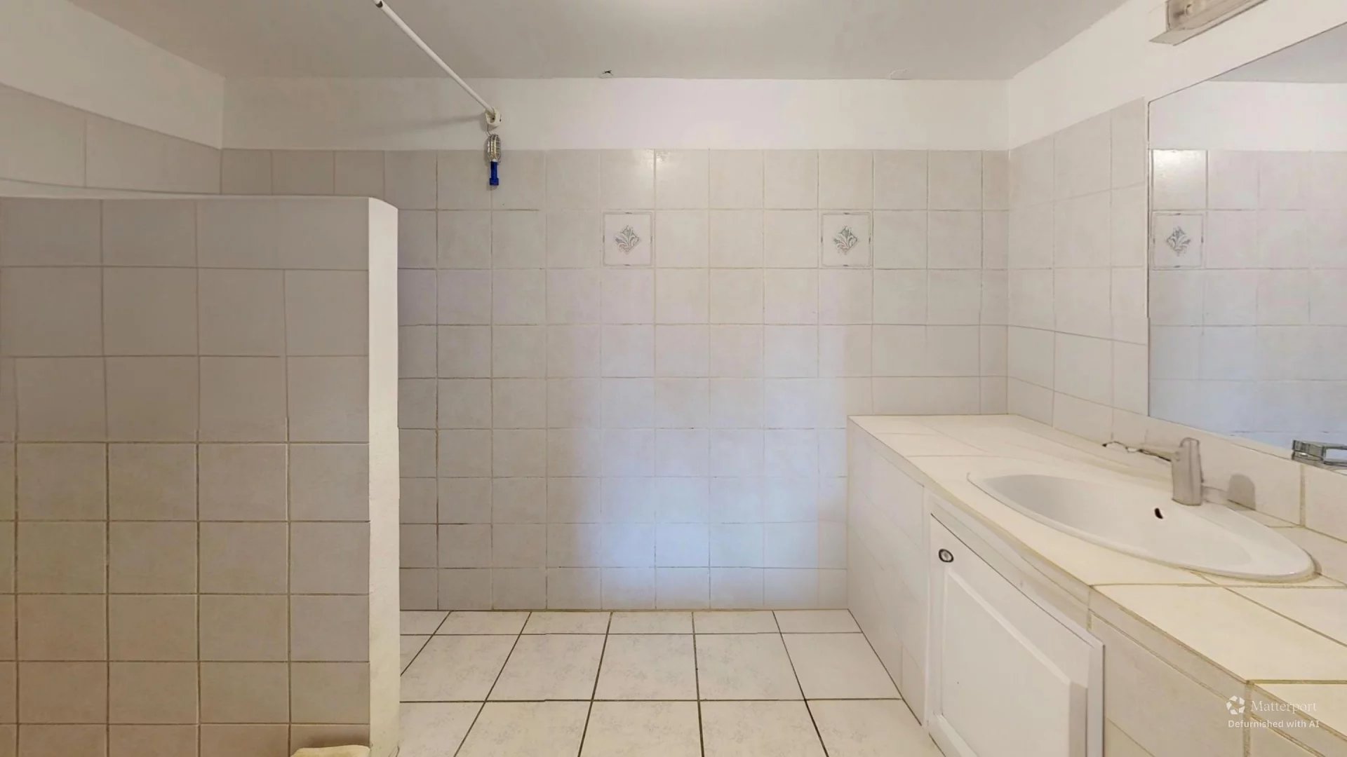 Empty white-tiled bathroom with a left-hand shower stall and a long white vanity with a sink on the right.