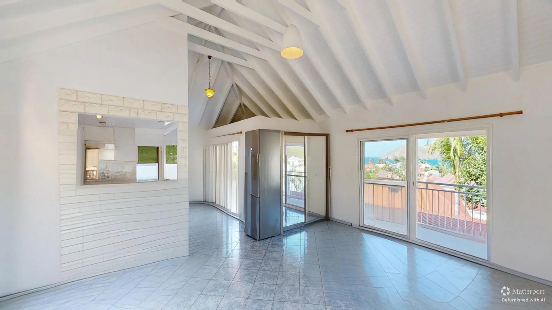 Bright open living space with vaulted white ceiling beams, large sliding doors to a balcony and ocean view, and a stainless steel fridge nearby.