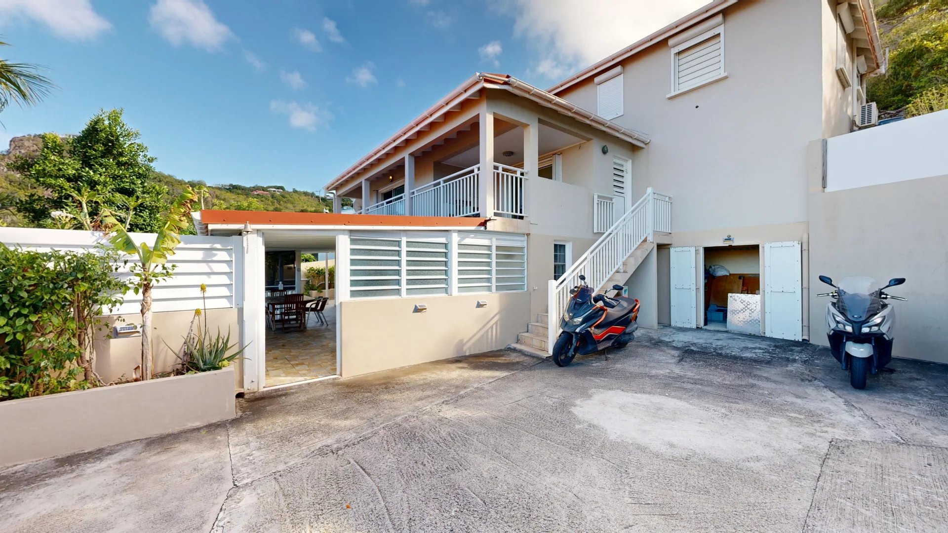 Two-story beige house with white railings, a covered patio, and stairs leading to the upper level, with two motorbikes parked nearby.