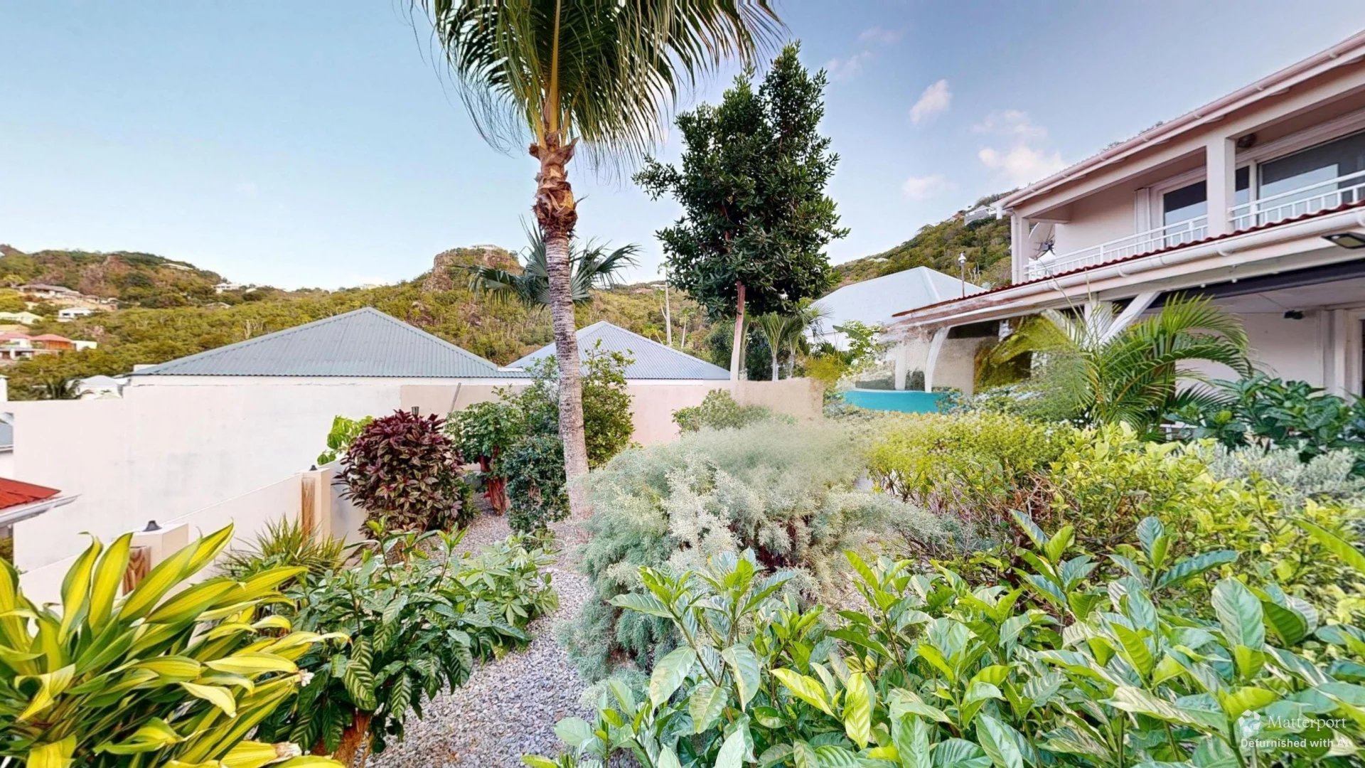 Lush tropical garden in a residential yard with a tall palm, assorted shrubs, and a white house with balconies.