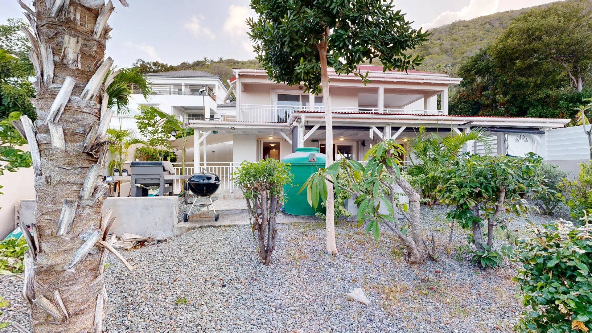 Backyard of a two‑story house with a gravel yard, tropical plants, and a green water tank beside a charcoal grill.