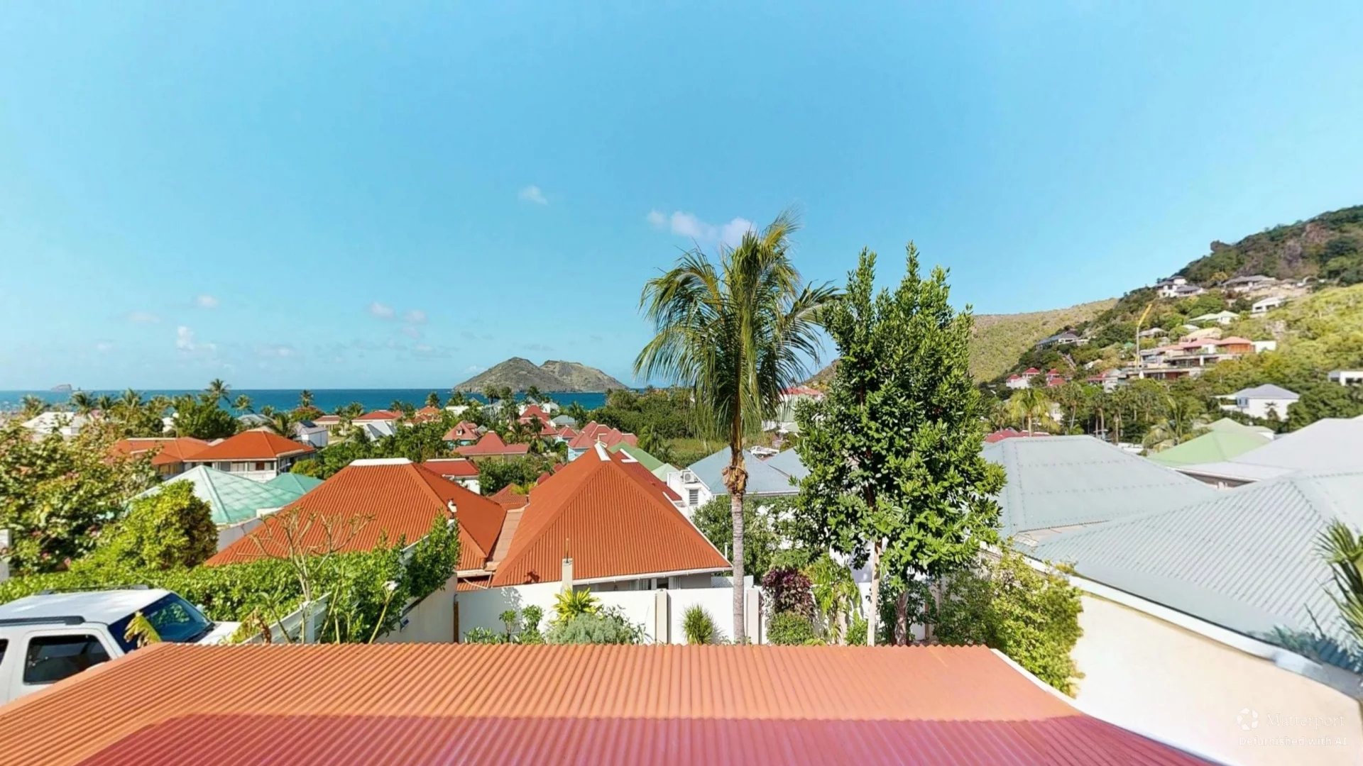 Aerial view of a tropical coastal town with red-tiled roofs, palm trees, and a blue sea in the distance.