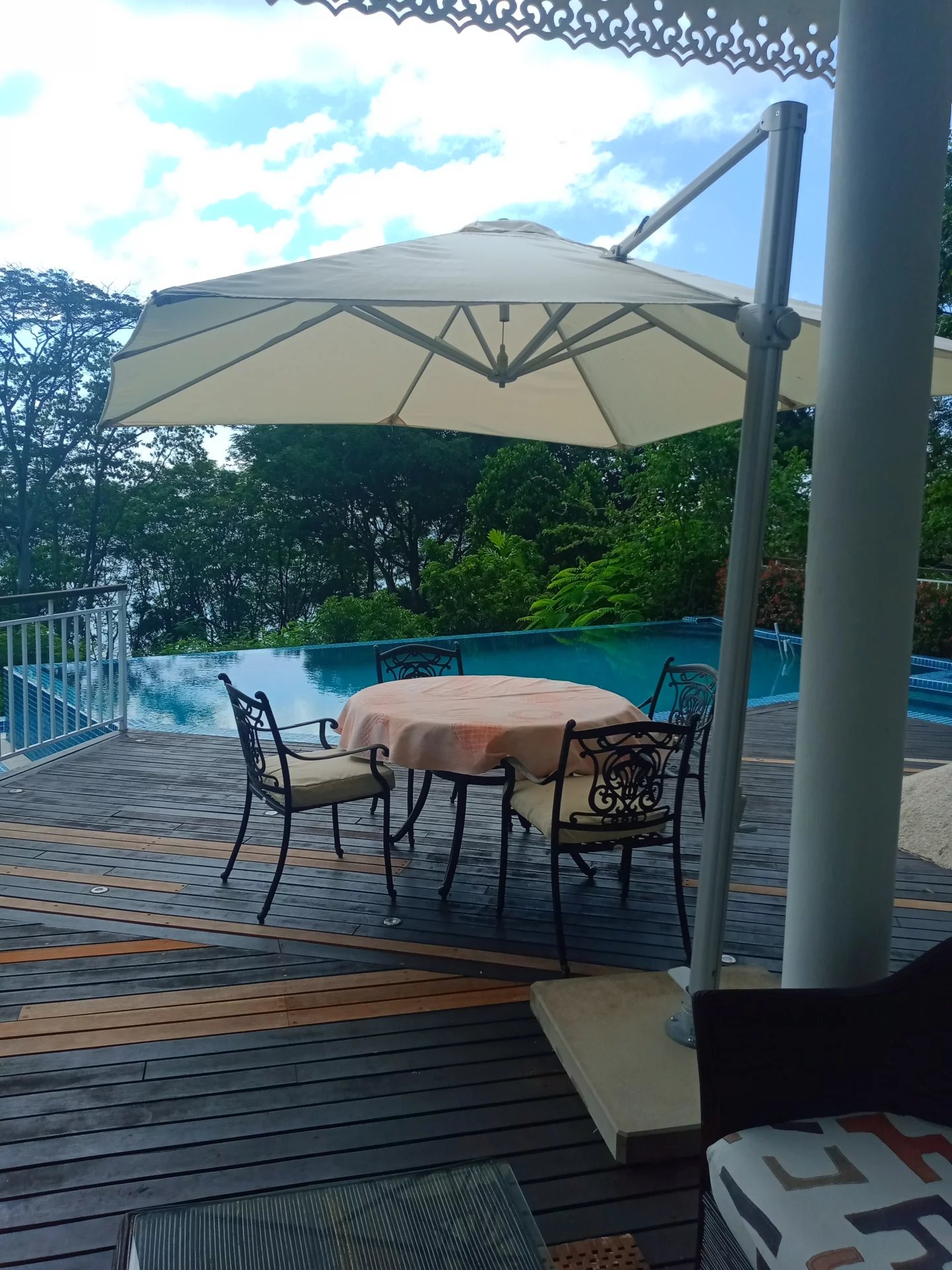 Round table with a peach tablecloth on a wooden deck, surrounded by four ornate metal chairs, under a large beige patio umbrella by a pool with greenery in the background.