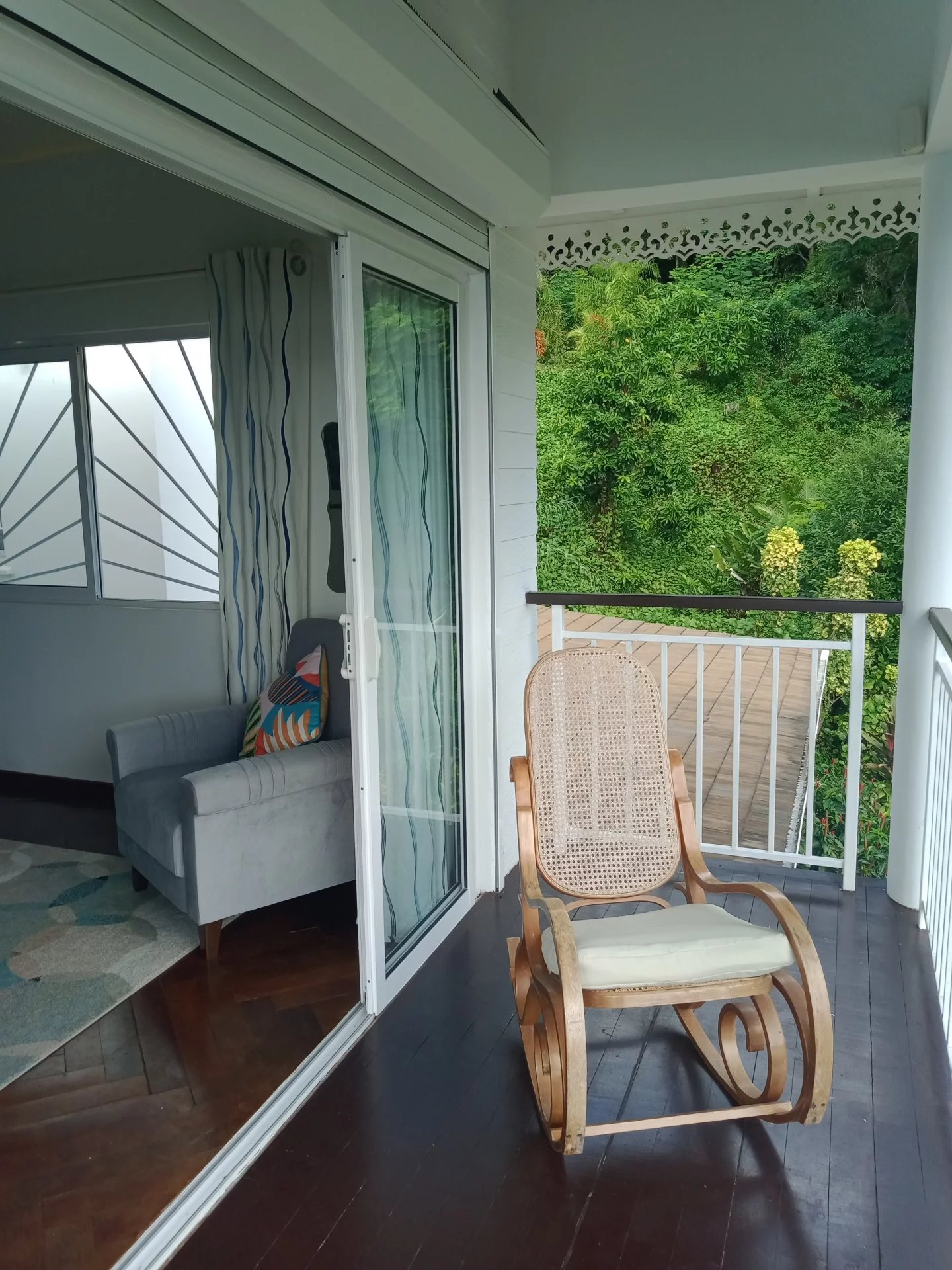 Wooden rocking chair on a dark deck, facing a lush green garden beyond a white railing. Sliding door to an interior with a gray sofa and blue curtains nearby.
