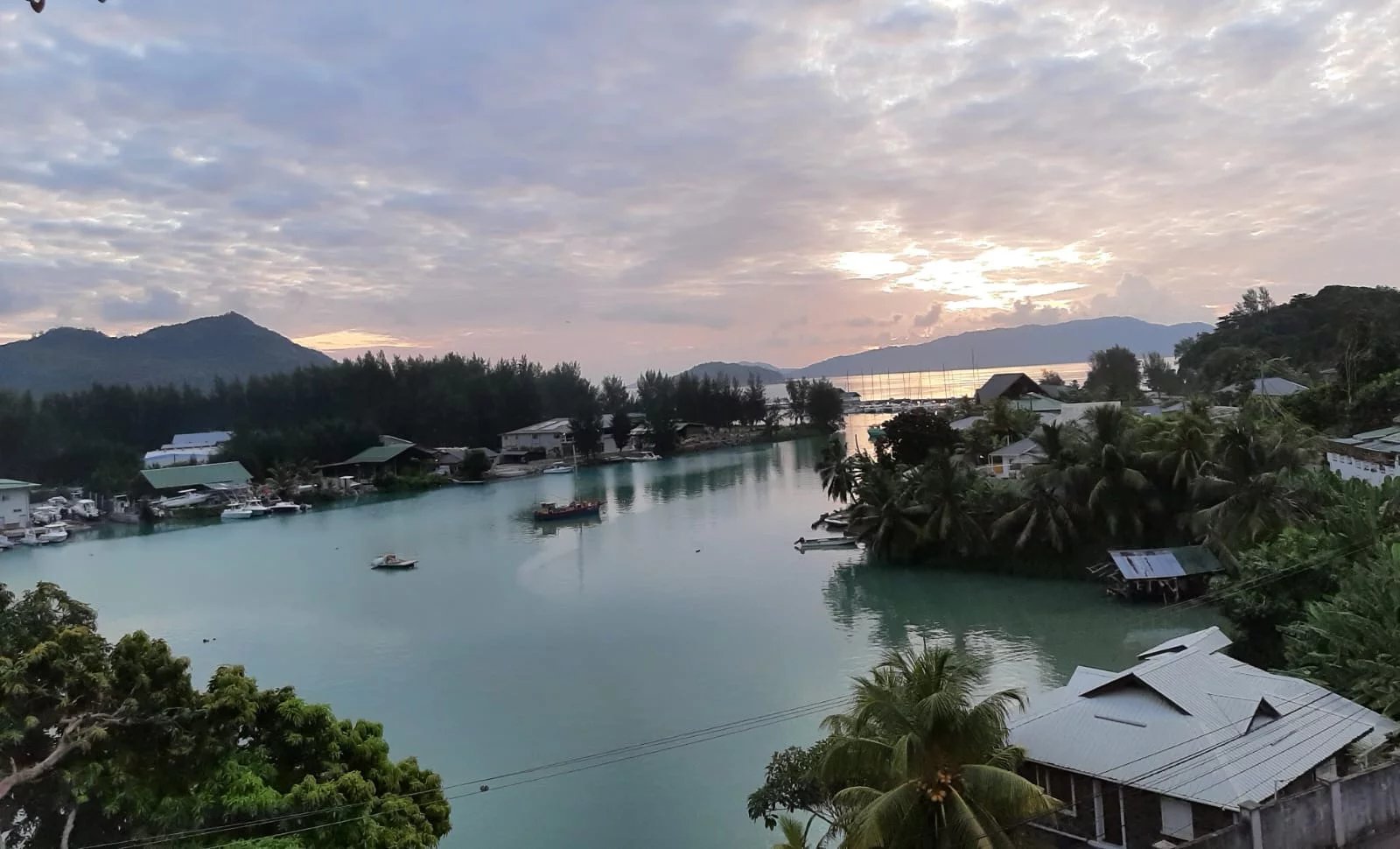 Sunset over a tropical harbor with turquoise water, boats, palm trees, and hills in the distance.