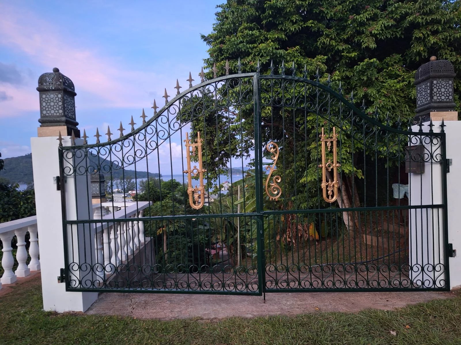 Green wrought-iron gate with gold ornamental accents, flanked by white pillars and lanterns, opening to a lawn with trees and a distant sea view