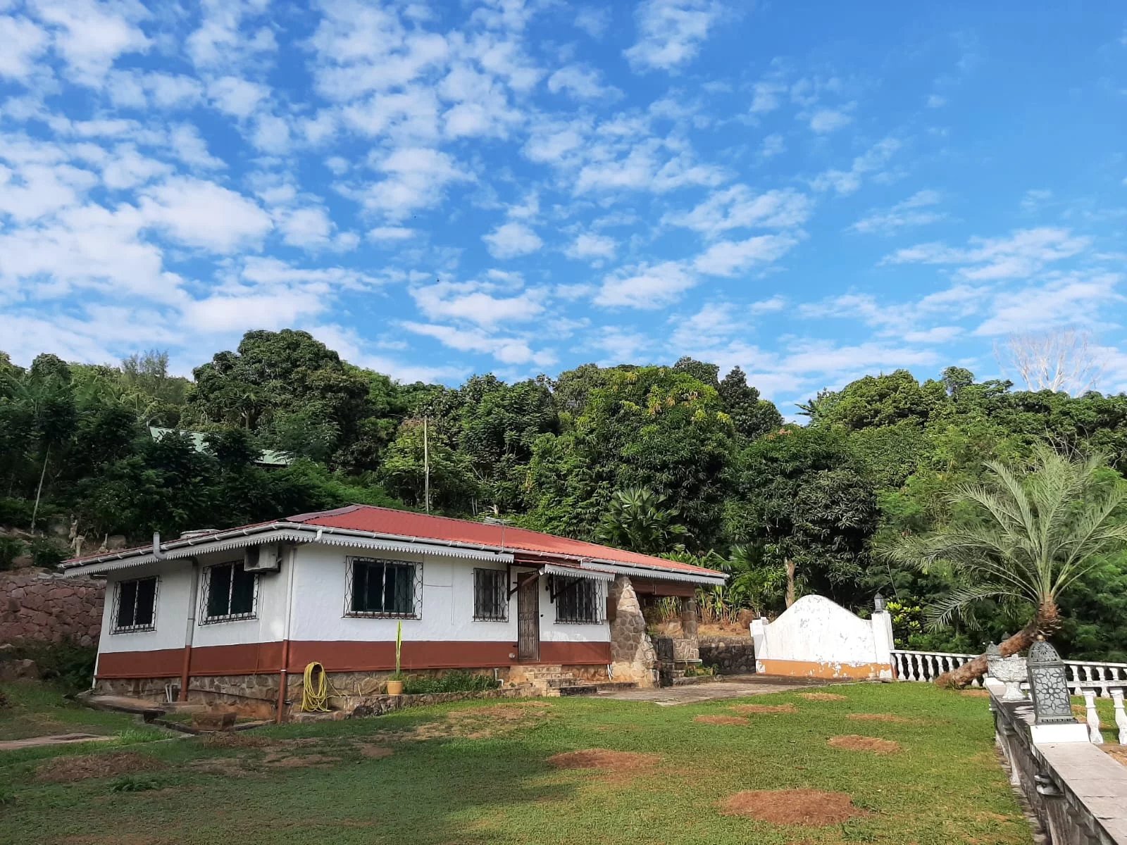 White cottage with a red roof sits on a grassy yard, surrounded by tropical trees under a bright blue sky with scattered clouds.