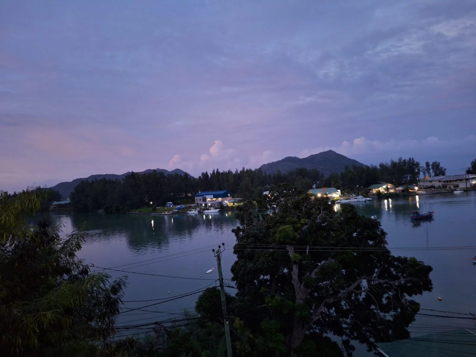 Calm harbor at dusk with boats along a tree-lined shore and distant mountains visible beyond the water's edge.
