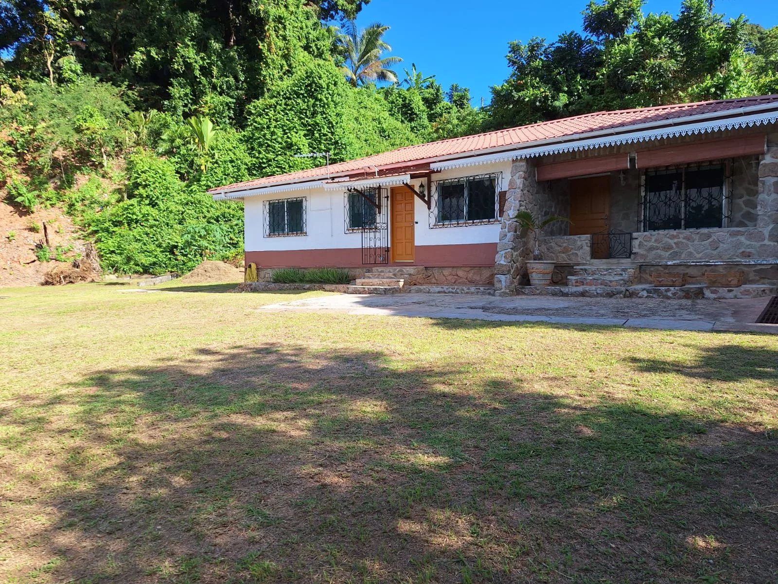 A single-story house with a white and stone exterior, red-tiled roof, and a front lawn under a sunny sky.