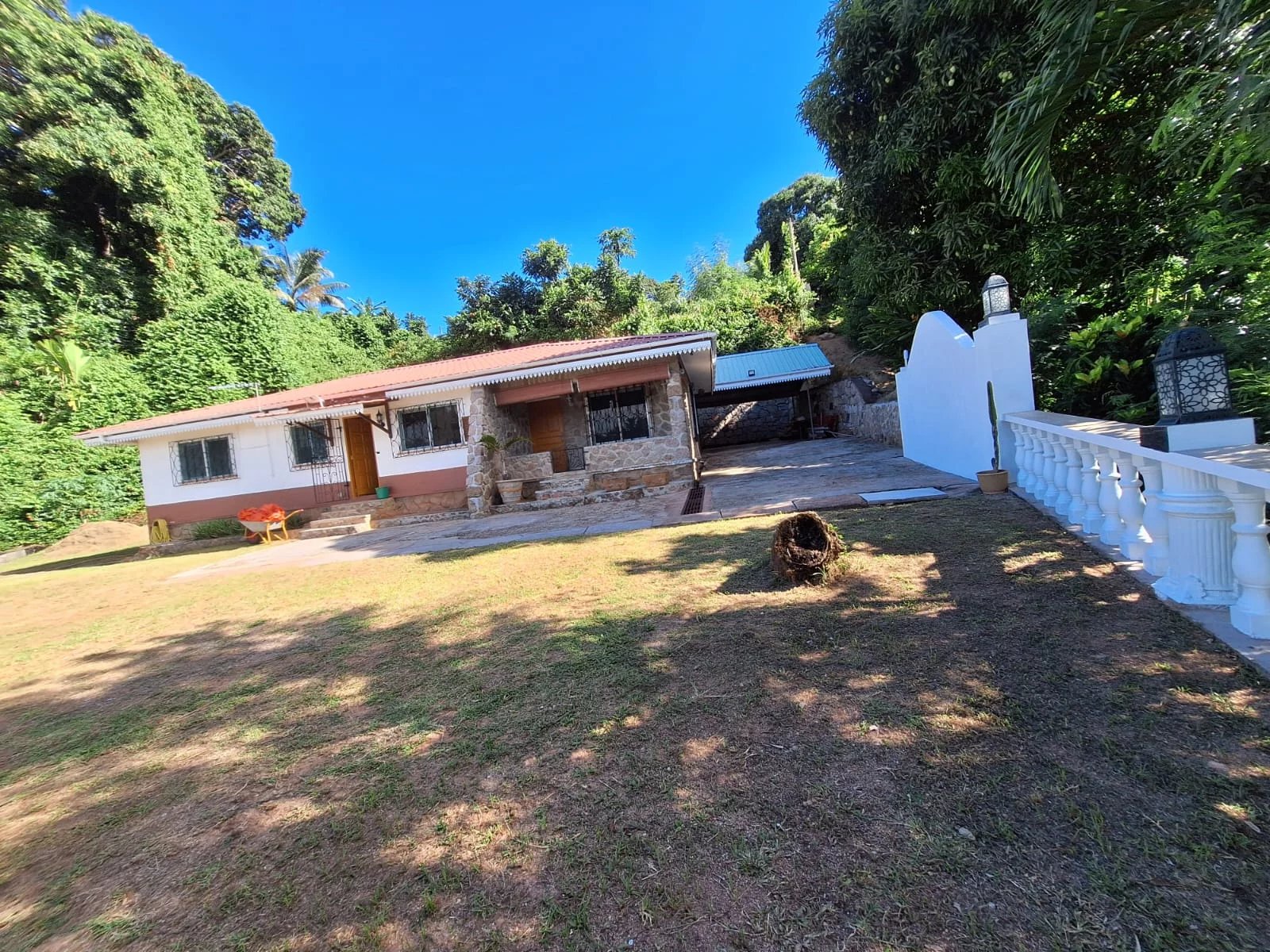 Single-story house with stone accents and a red-tiled roof, in a sunny yard with trees and a blue sky.