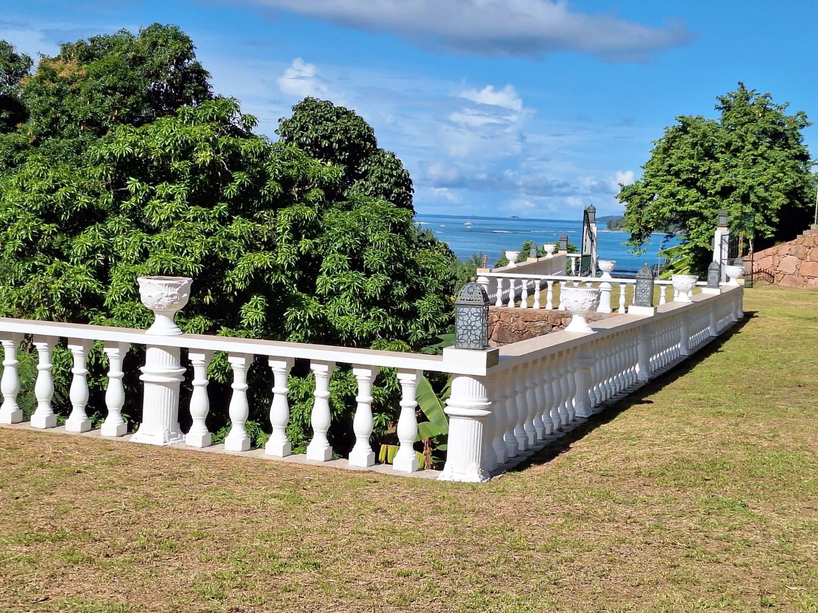 White ornate balustrade terrace with urns, overlooking the blue sea and green trees under a bright sky.