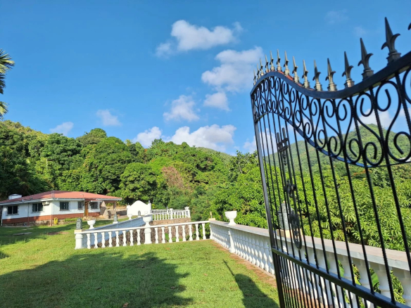 Ornate wrought iron gate on the right opening to a sunny yard with a white balustrade green lawn and a red roofed house beyond Propriétés clovis