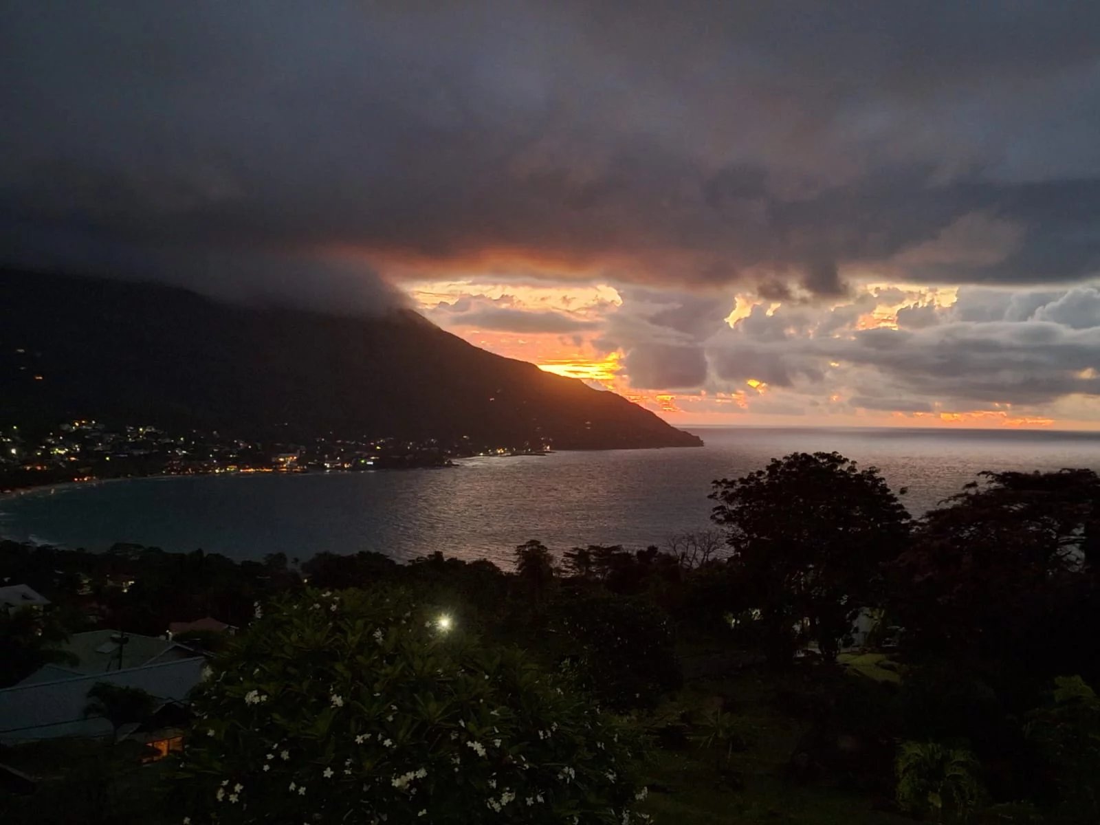 Coastline at sunset with dark clouds over a hilly cape, calm sea reflecting orange light, and lights of a town along the shore in the foreground trees and flowers in silhouette.