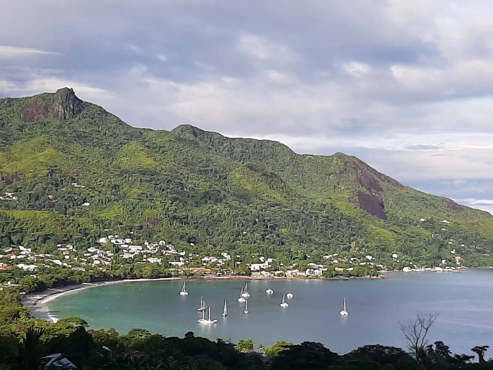 Tropical bay with a crescent sandy beach and calm turquoise water, small white houses along the shore, and forested green hills in the background under a cloudy sky.