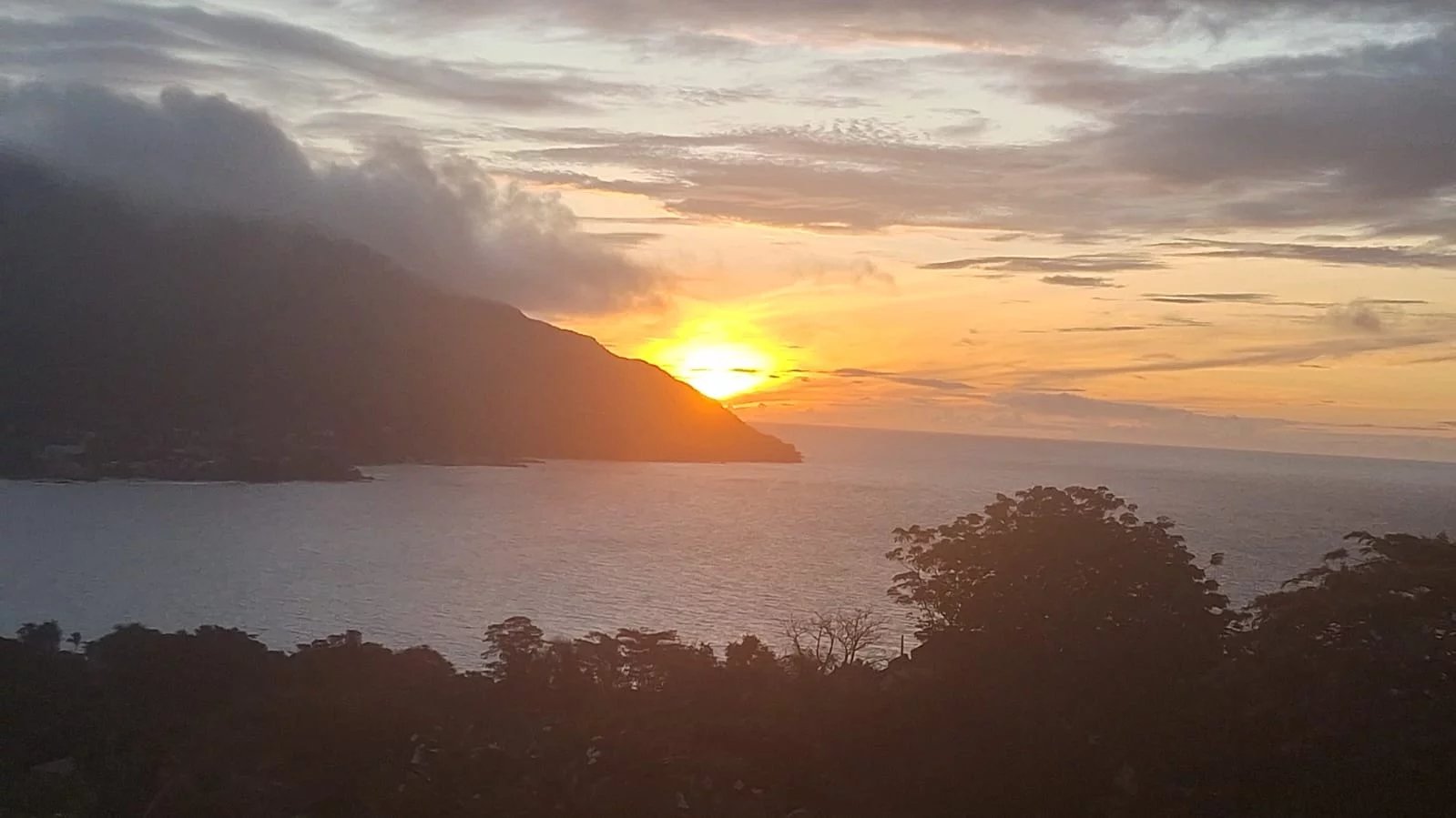 Sunset over calm sea with a silhouetted hillside on the left and trees in the foreground.