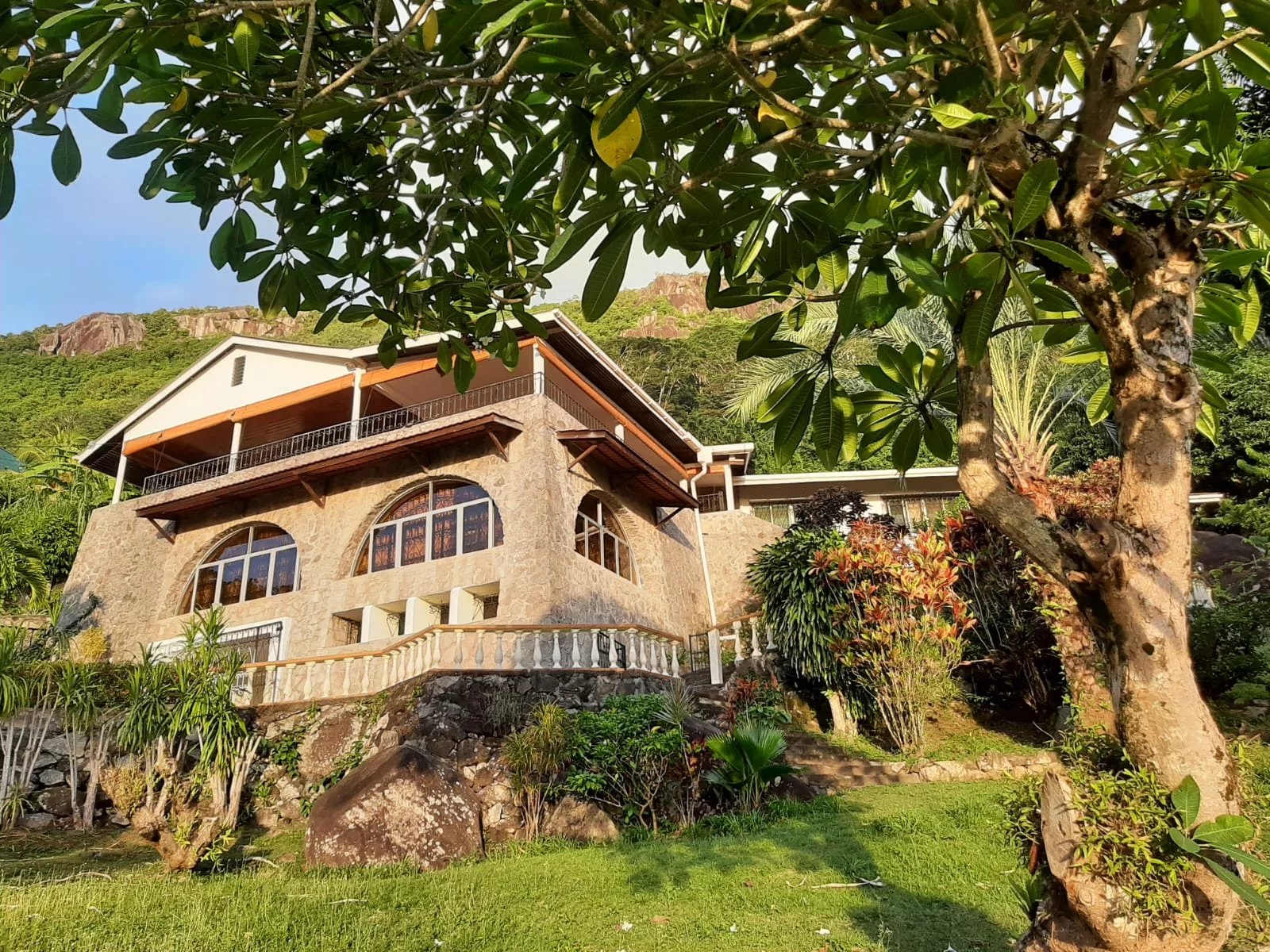 Stone hillside villa with arched windows, wooden balcony, and lush tropical garden surrounding it.
