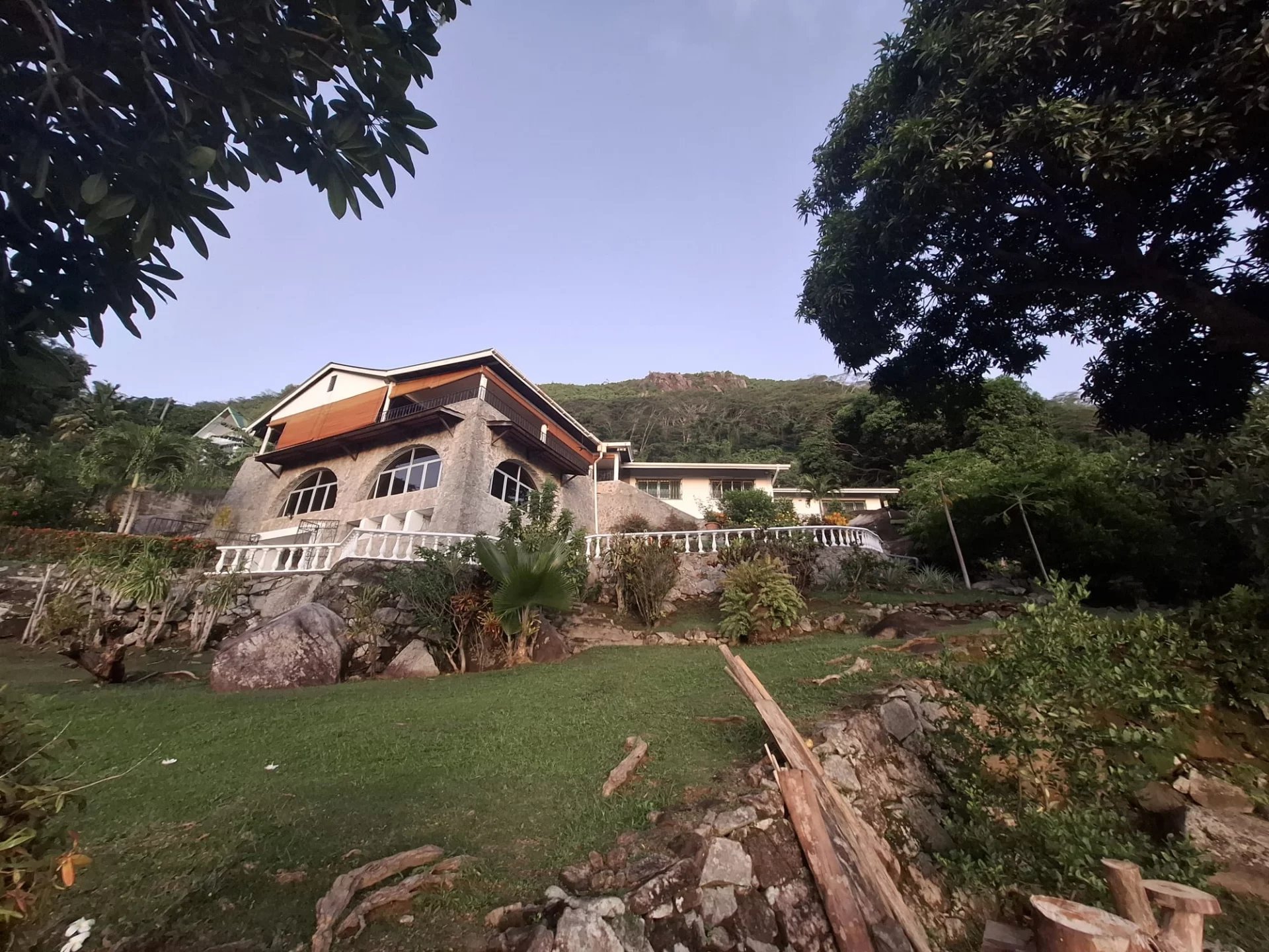 Stone hillside villa with arched windows and white balcony railing, surrounded by tropical garden under a clear blue sky.