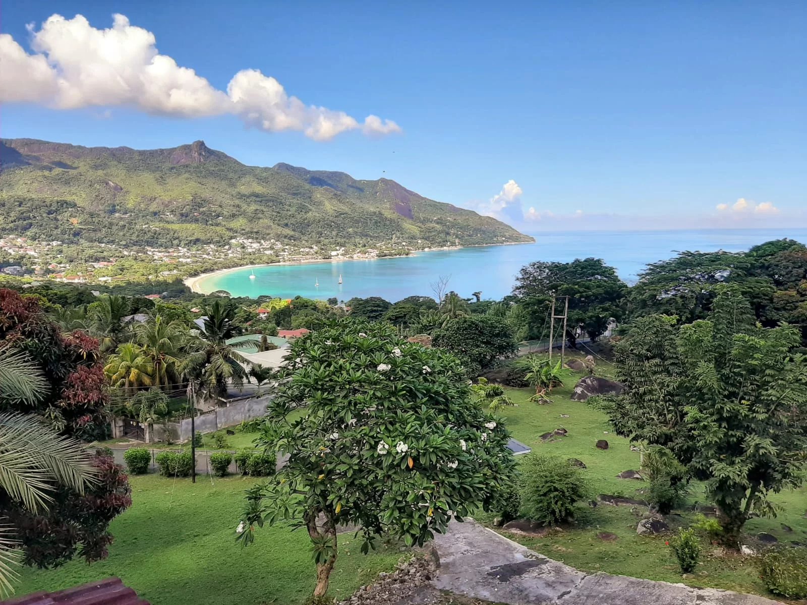 Tropical coastal landscape: turquoise bay curving along a green hillside with a village at the shore and blue sky above.