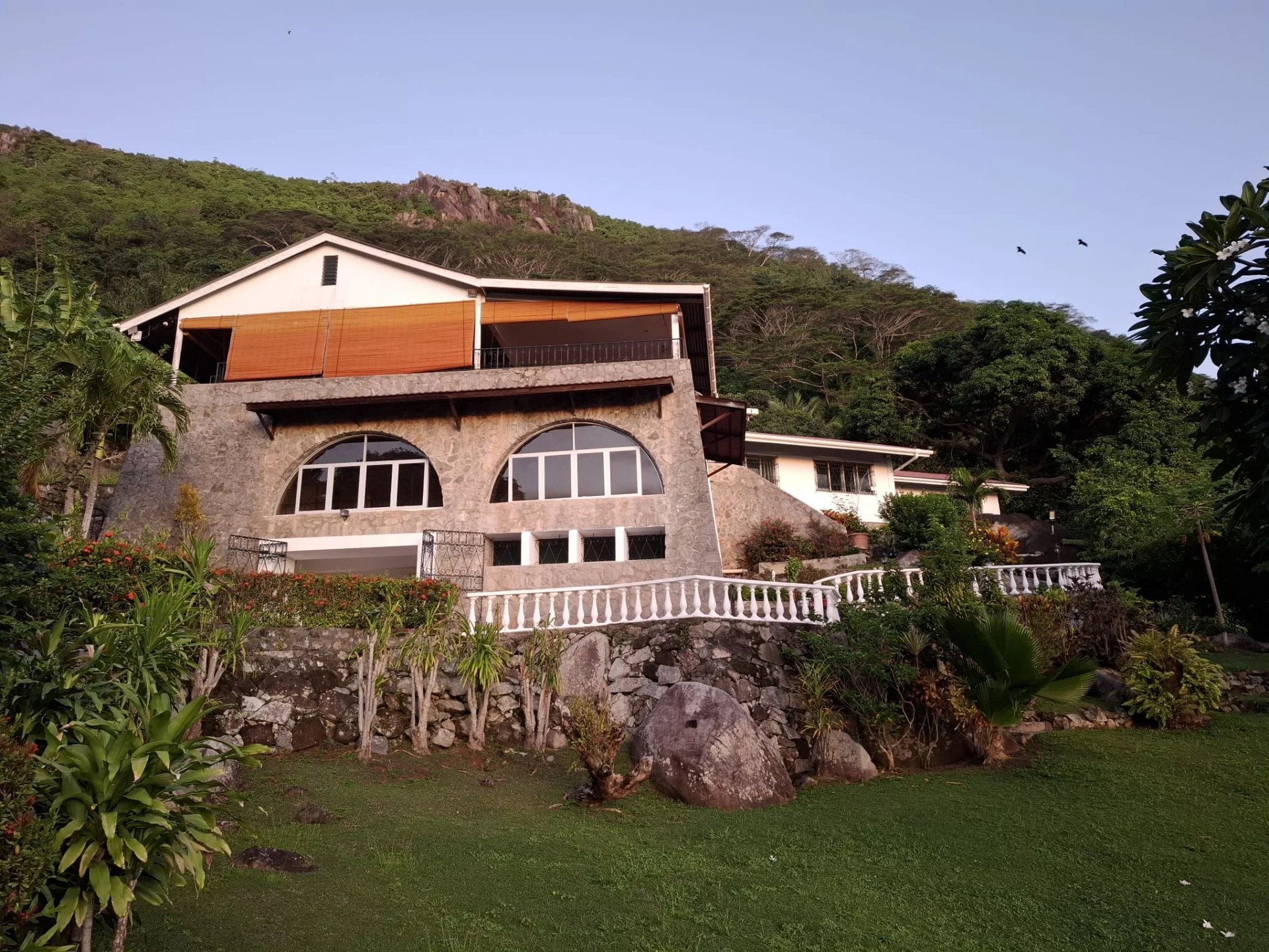 Stone hillside villa with arched windows and a white railing, set among tropical plants and rocks on a terraced garden.