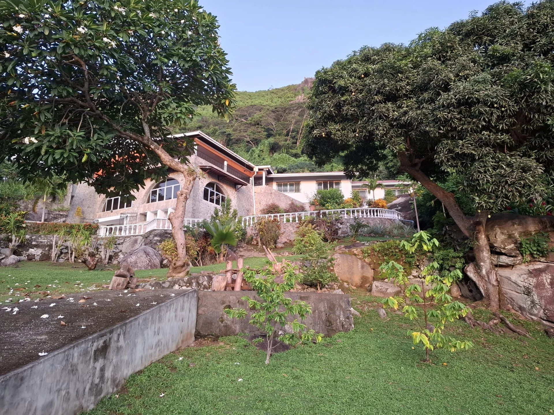 Stone house with arched windows and a white railing set into a landscaped garden, large trees framing the scene under a clear blue sky.