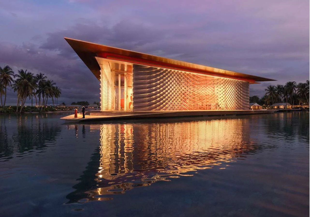 Floating pavilion on a calm lagoon, warmly lit with a copper roof and wavy metal walls at dusk, palm trees in the background.