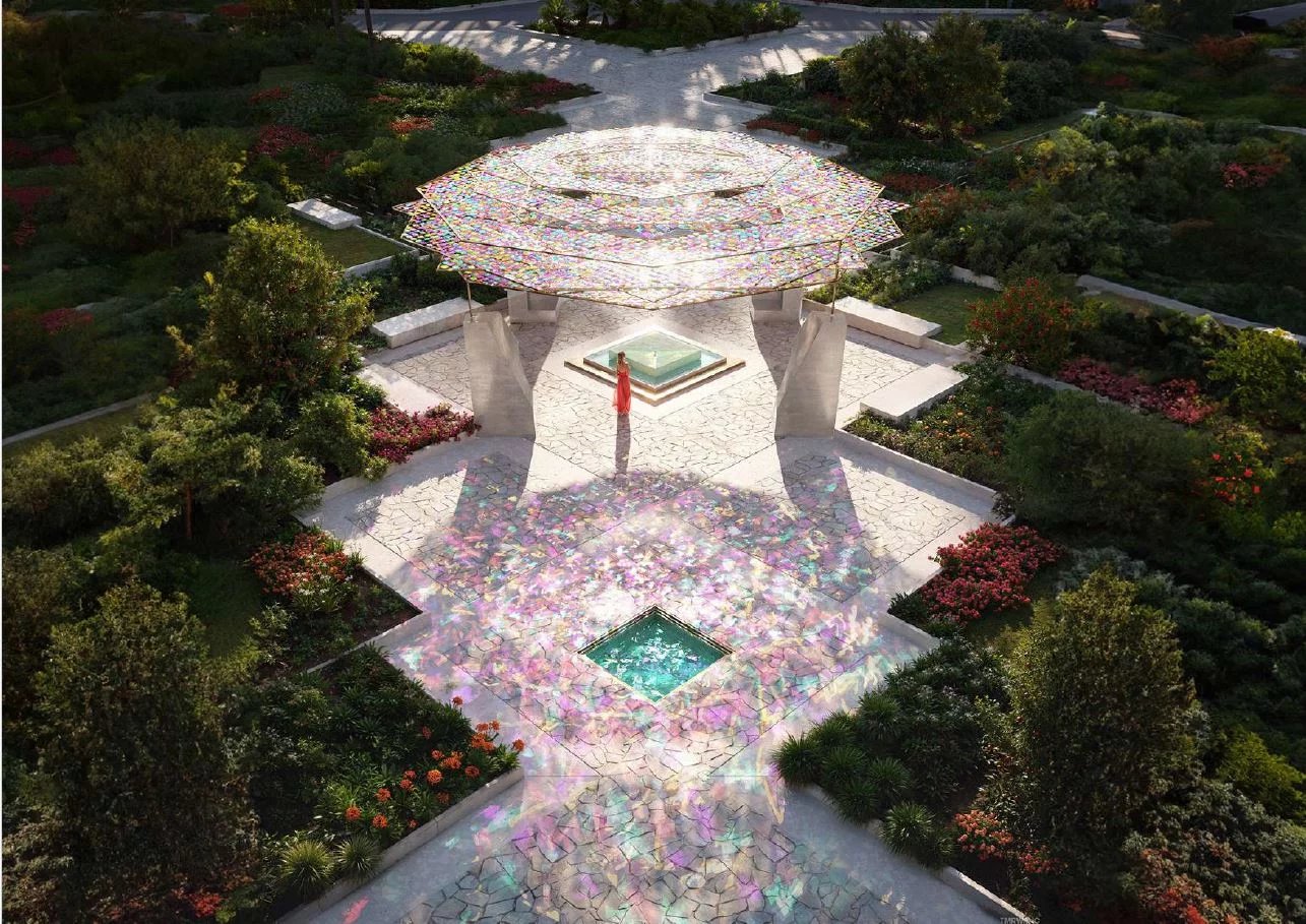 Aerial view of a garden with a colorful mosaic canopy over a stone pavilion and a person in a red dress standing near a central fountain.