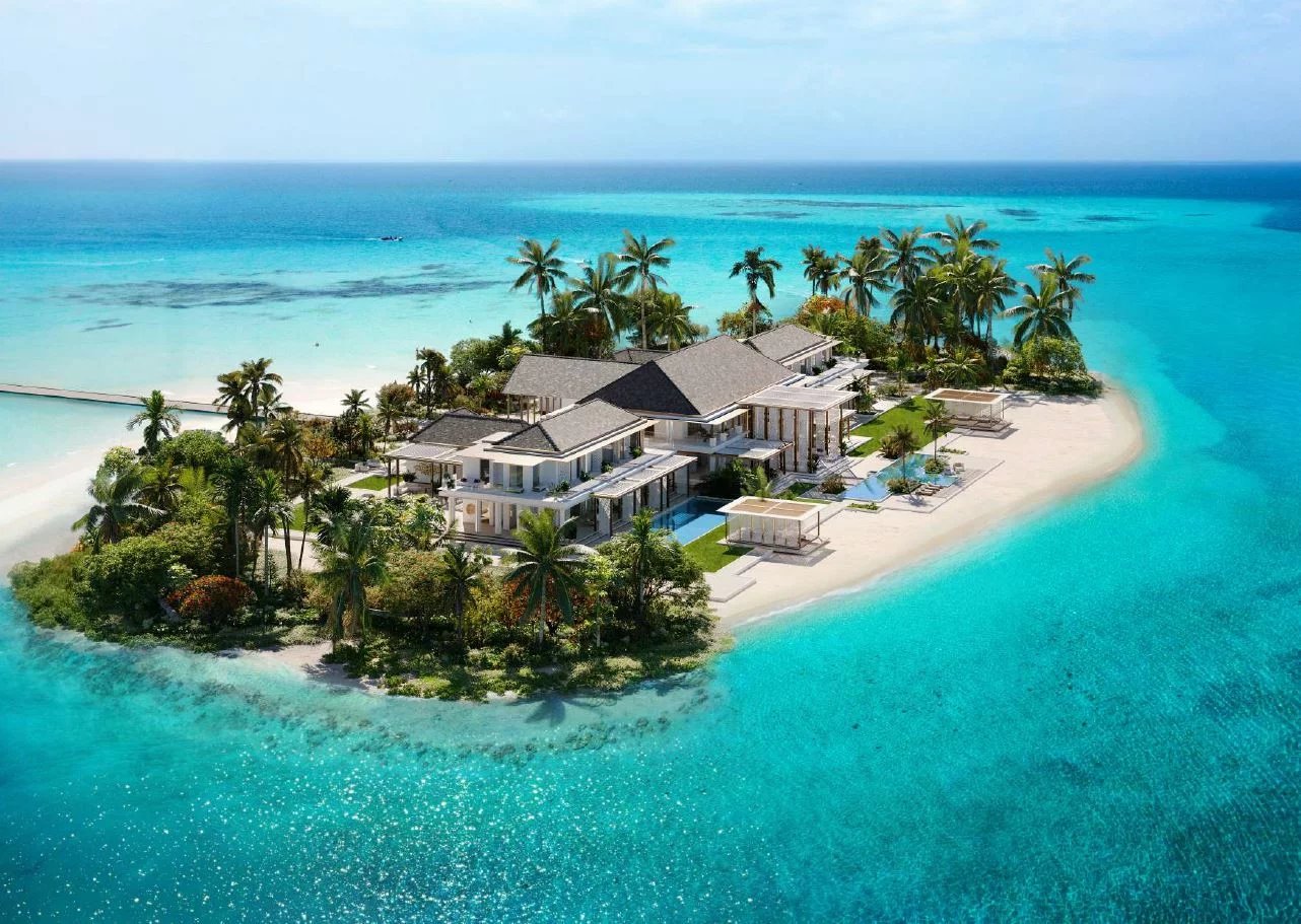 Luxurious beachfront villa complex on a small tropical island amid turquoise shallow waters and palm trees.