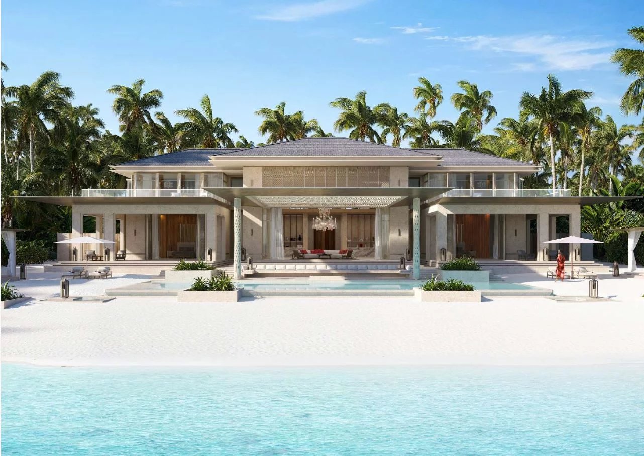 Luxurious beachfront villa with a spacious pool, white sand, and palm trees under a clear blue sky.