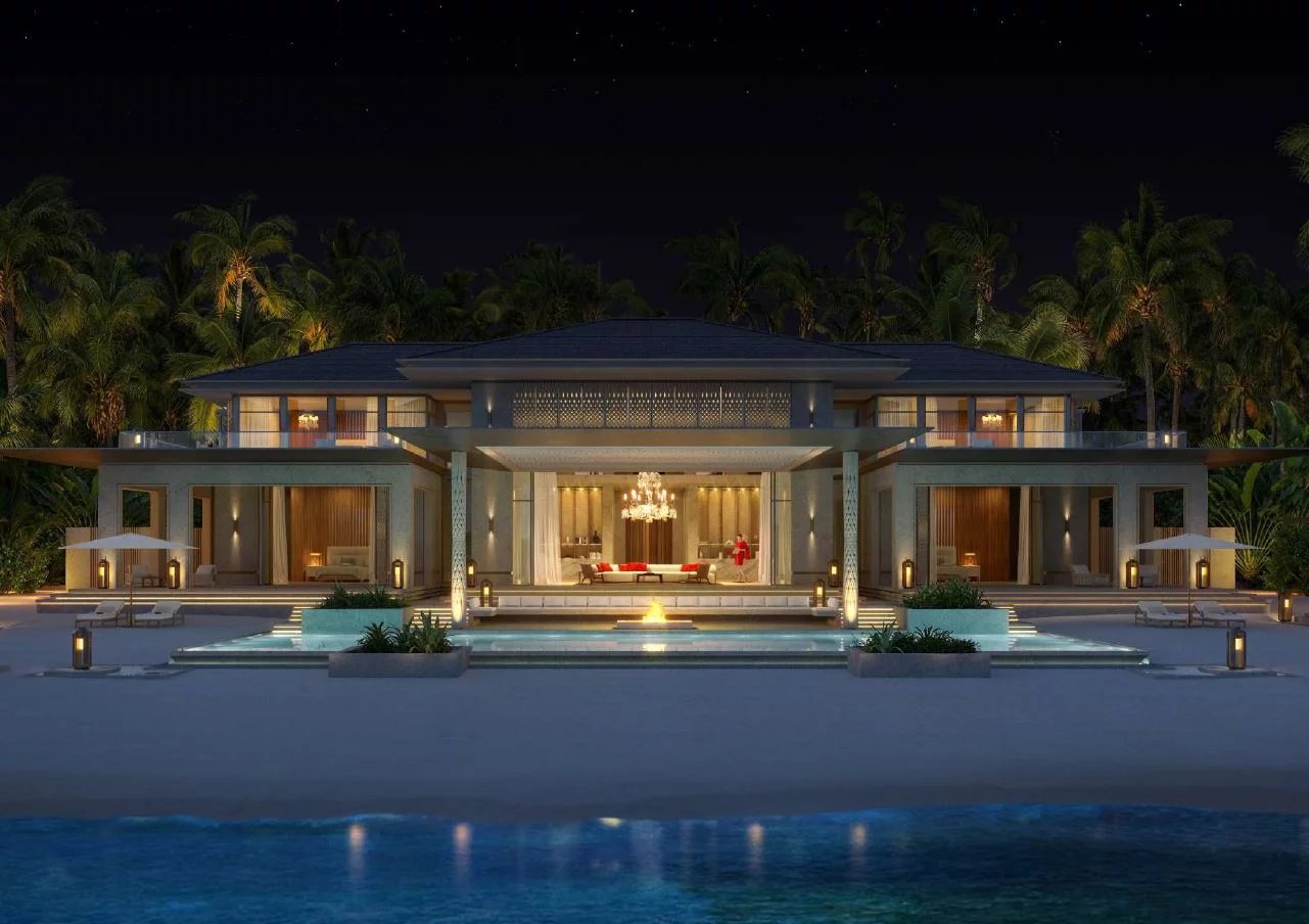 Luxurious beachfront villa at night with a lit central fountain, pool, and palm trees surrounding the modern two-story residence.