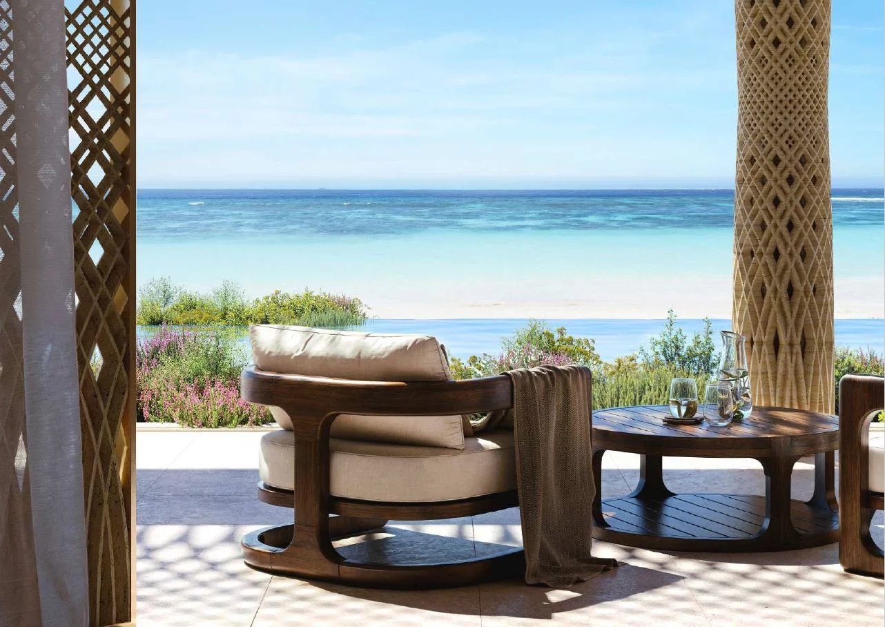 Patio seating area with beige cushioned chair and round wooden table overlooking a turquoise ocean and white sand beach.
