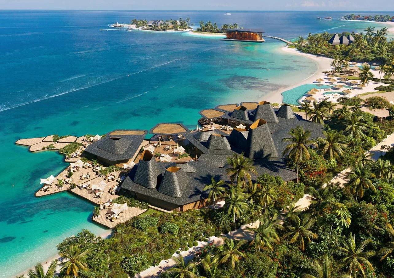 Aerial view of a tropical island resort with dark thatched roofs, palm trees, and turquoise lagoon waters surrounding wooden decks and white-sand beaches.