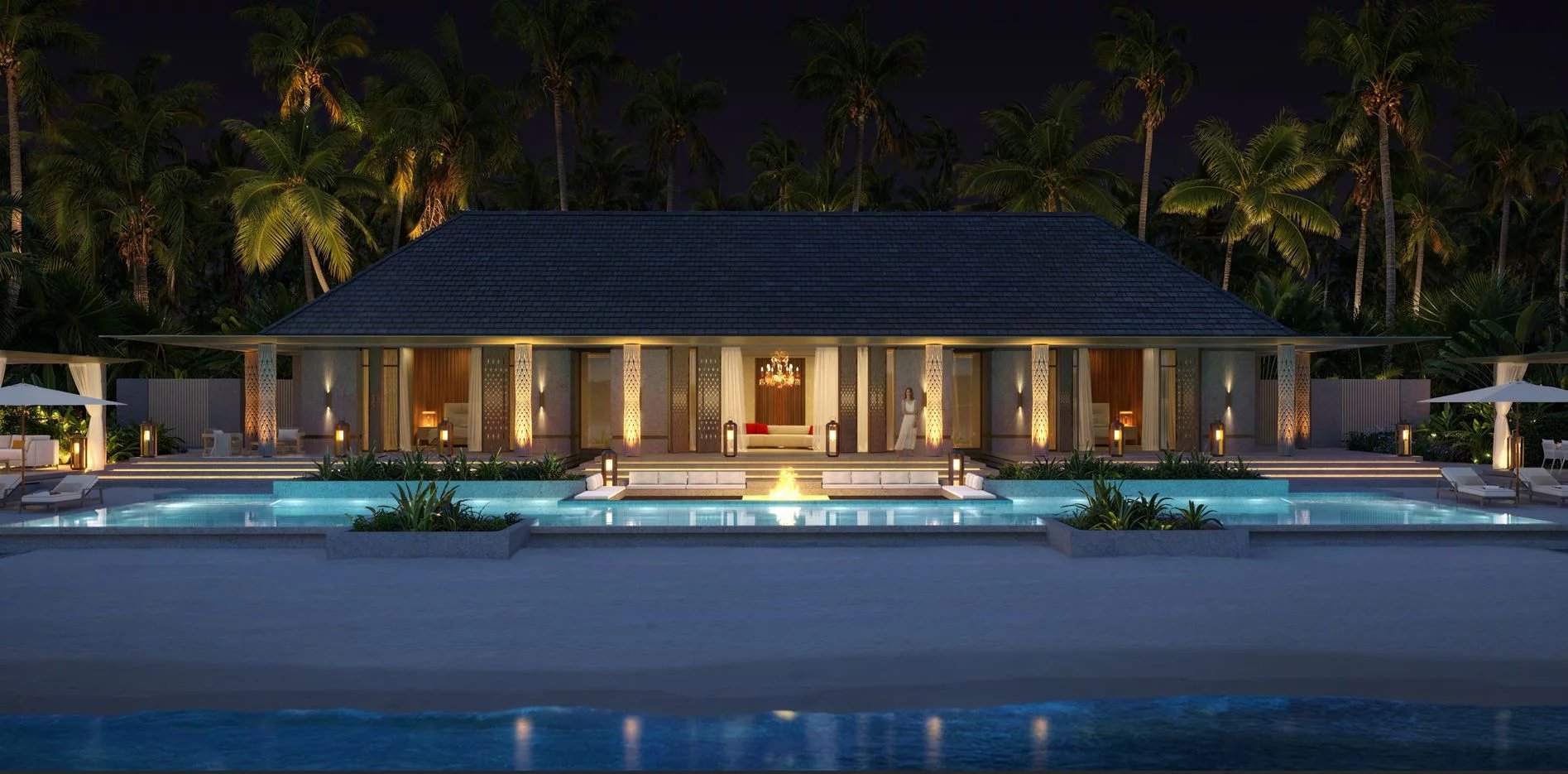 Luxurious beachfront villa at night with illuminated courtyard, central fire pit, and two-tier swimming pools surrounded by palm trees and lounge areas