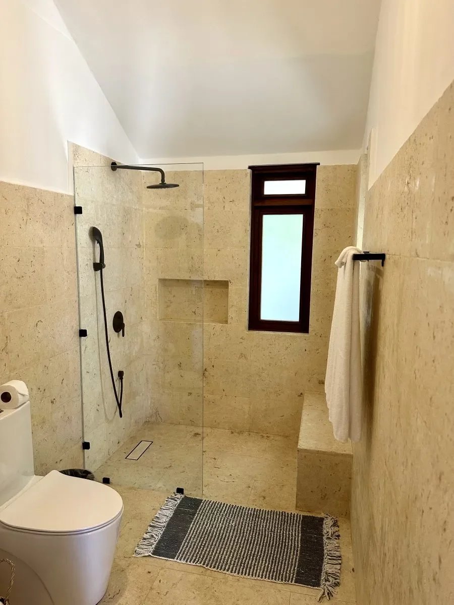 Modern bathroom with a glass-enclosed walk-in shower, beige tiled walls, and a white toilet nearby; a window and towel rack on the wall, plus a striped rug on the floor.