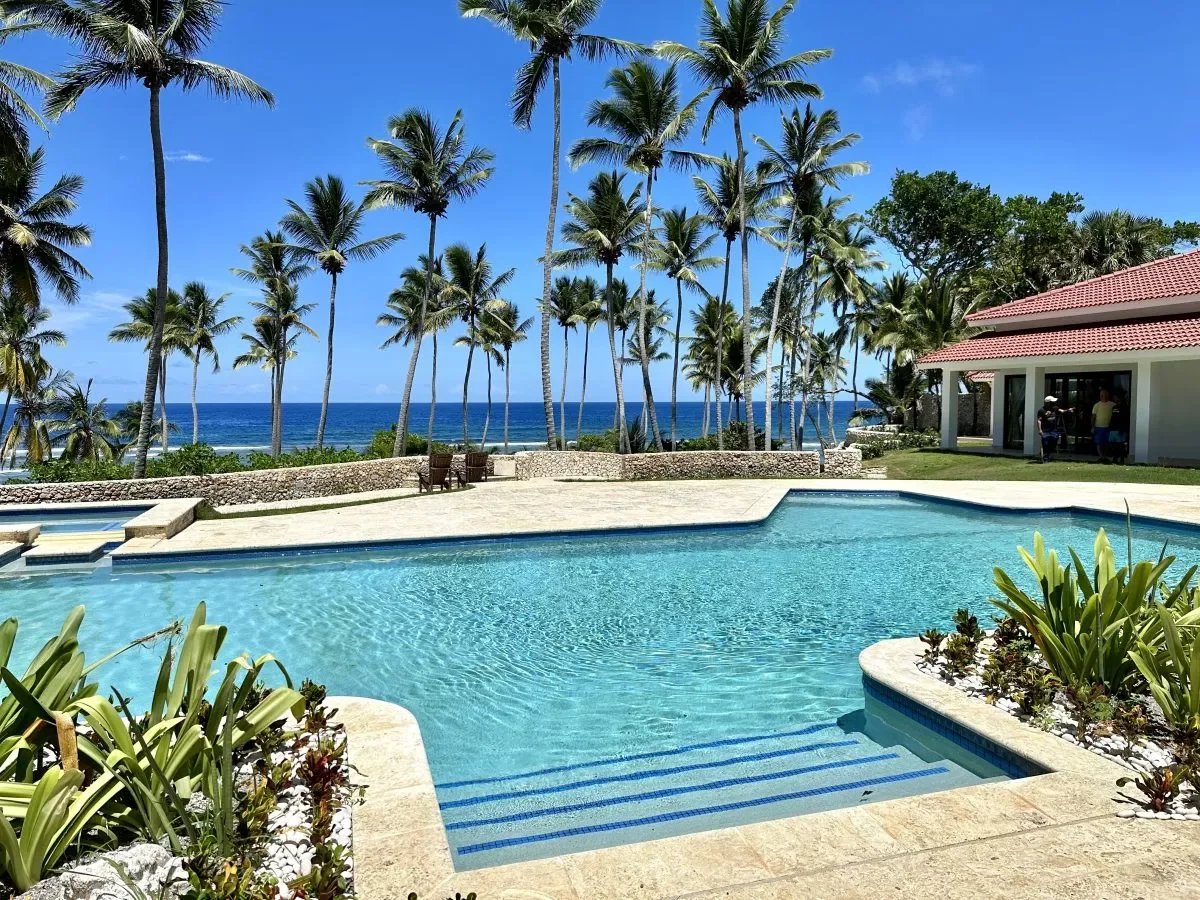 Poolside at a tropical resort with palm trees, a clear blue pool, and the ocean beyond under a bright sky and red-roofed building to the right.