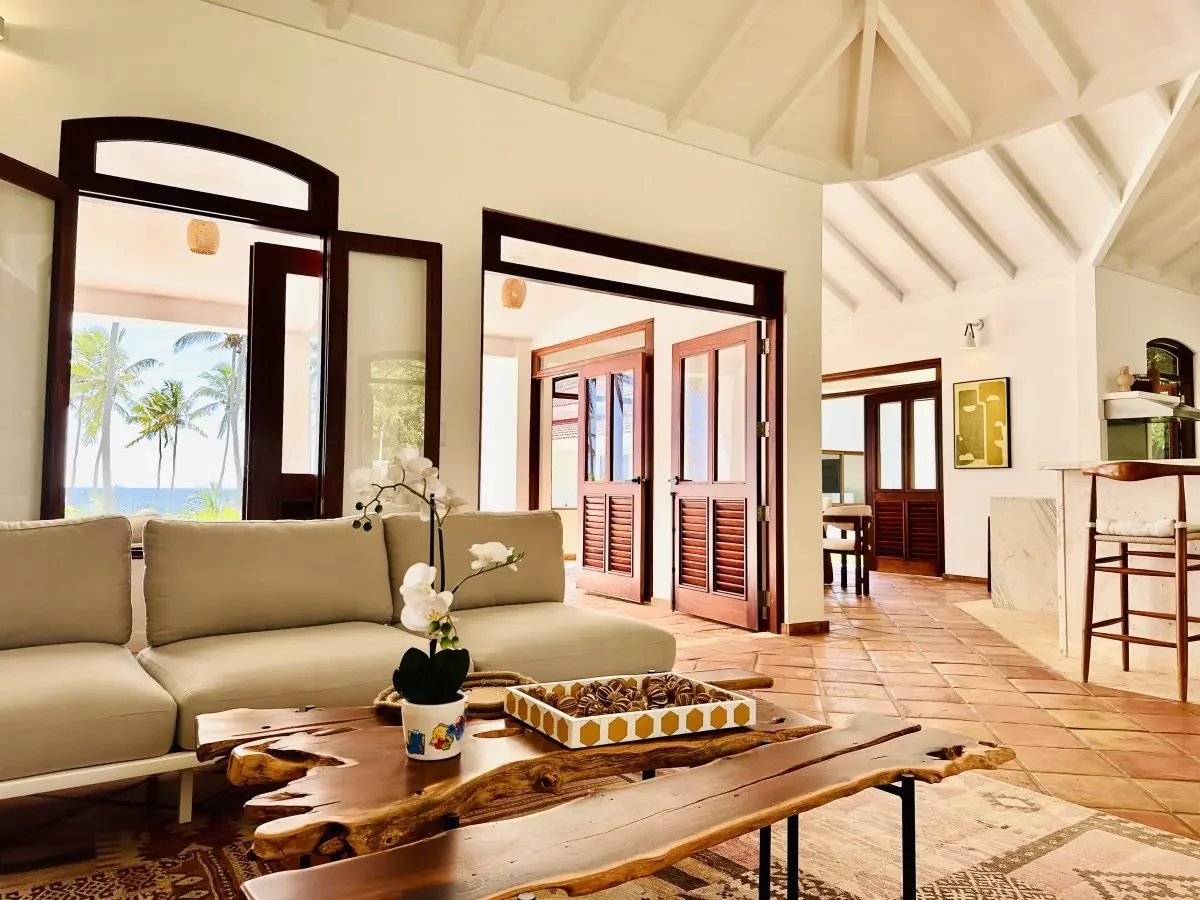 Bright tropical living room with white sofas, a natural wood coffee table, and open wooden doors leading to palm trees and the ocean outside.