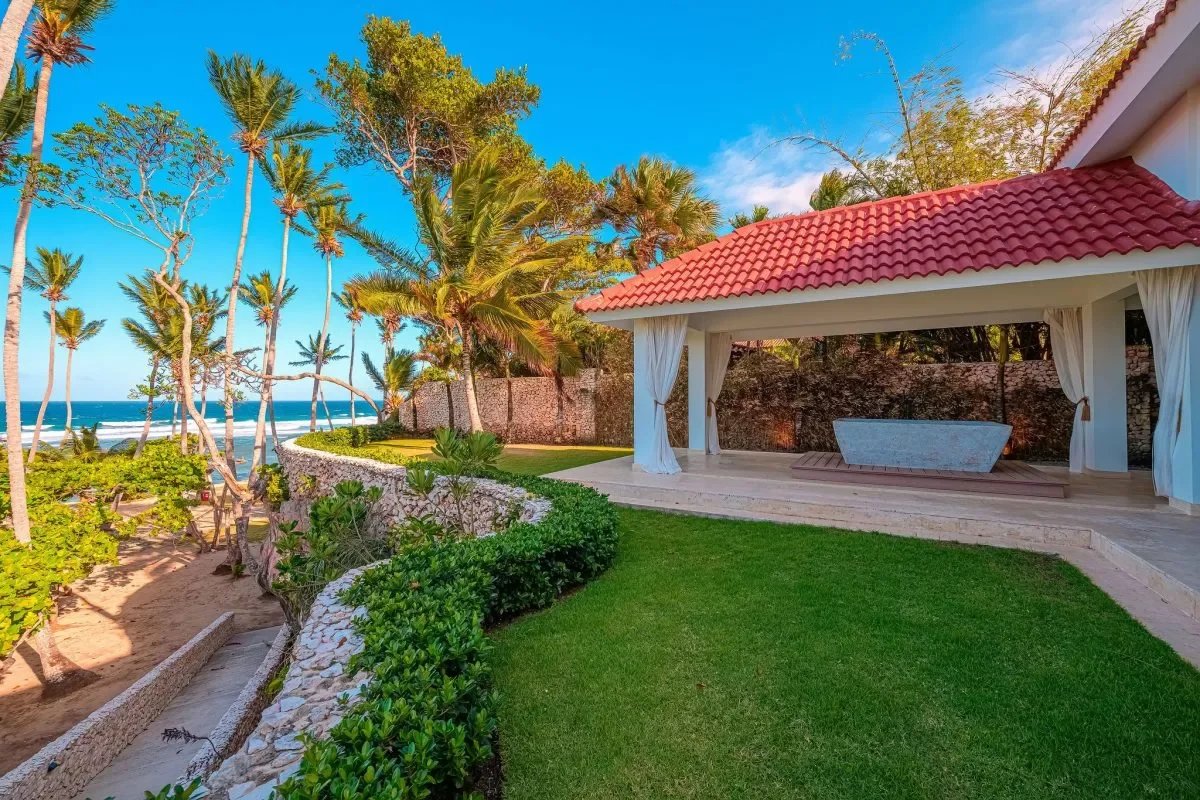 Beachfront gazebo with a red-tiled roof, white curtains, and a stone deck overlooking the ocean amid palm trees and green lawn.