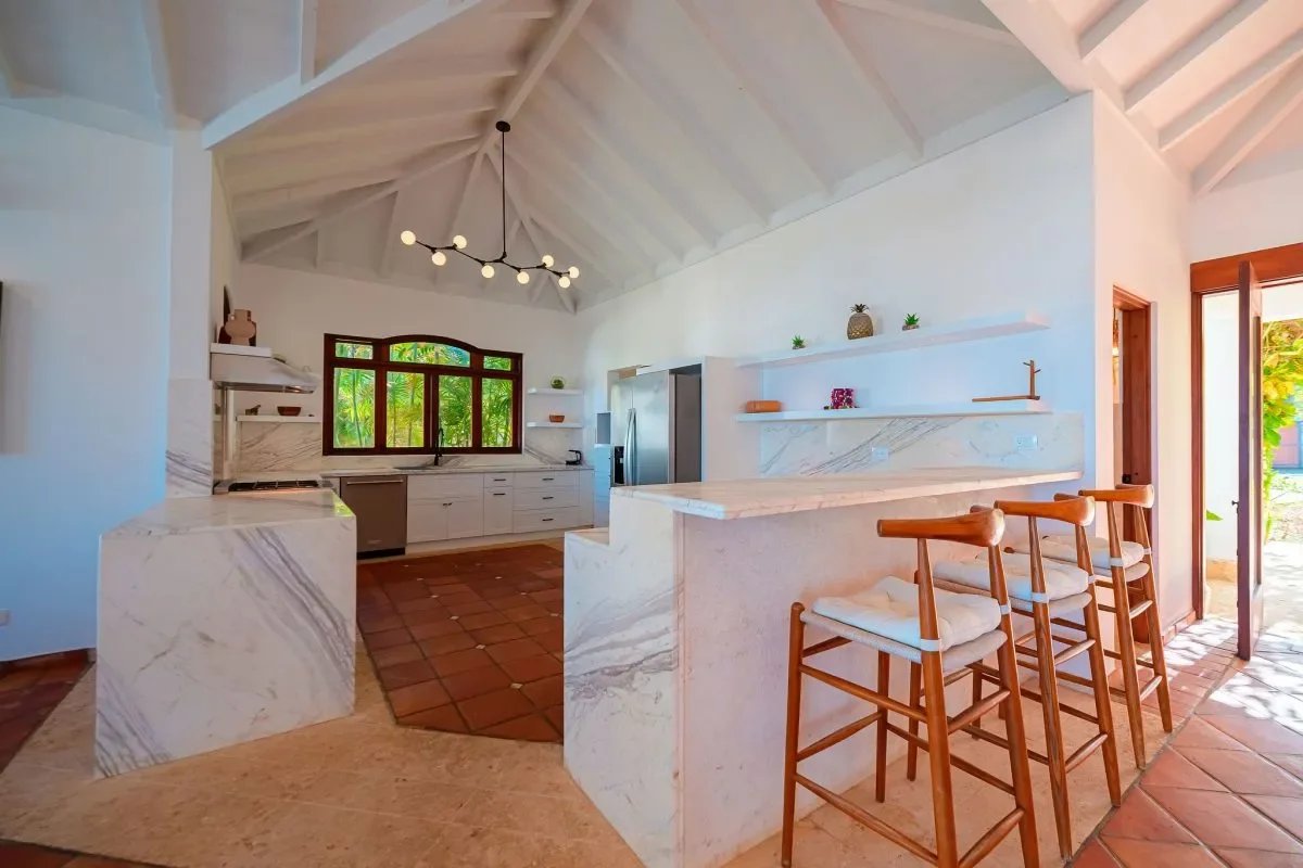 Bright open-concept kitchen with marble countertops, island, and wooden bar stools under a vaulted white ceiling; windowed door to a sunny patio.